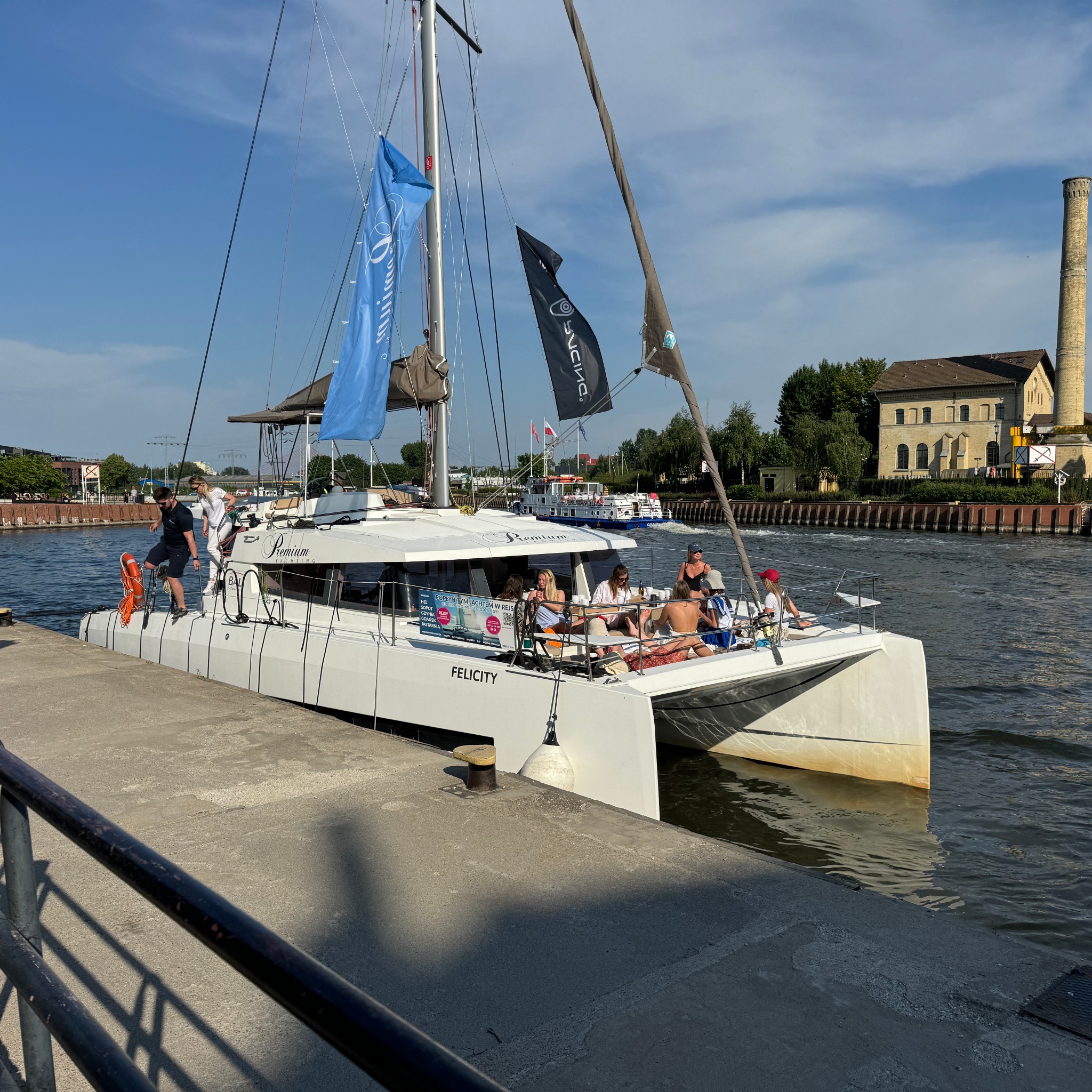 Biały katamaran 'Felicity' zacumowany przy nabrzeżu, z grupą pasażerów na pokładzie, widoczne flagi firmowe i budynek z kominem w tle.
