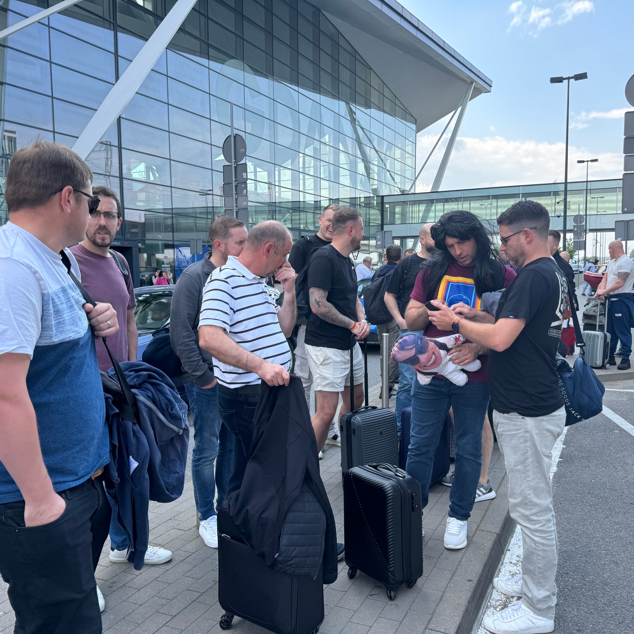 Grupa osób z bagażami przed nowoczesnym budynkiem lotniska; jeden mężczyzna w peruce trzyma pluszową poduszkę z nadrukiem twarzy, inny sprawdza coś na telefonie.