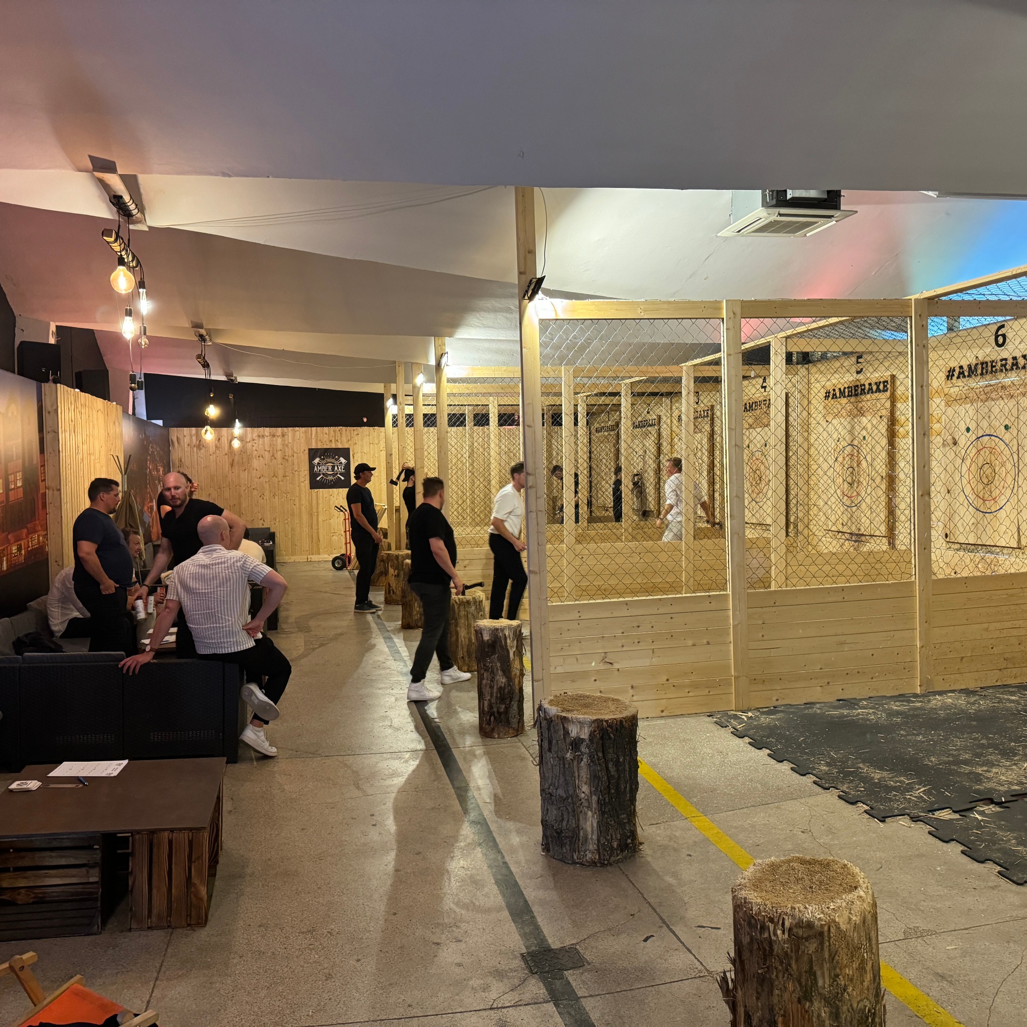 Wnętrze hali z torami do rzucania siekierami, grupa mężczyzn odpoczywa na sofie, a inni przygotowują się do rzutu, widoczne drewniane tarcze z numerami i hashtagiem #AMBERAXE.