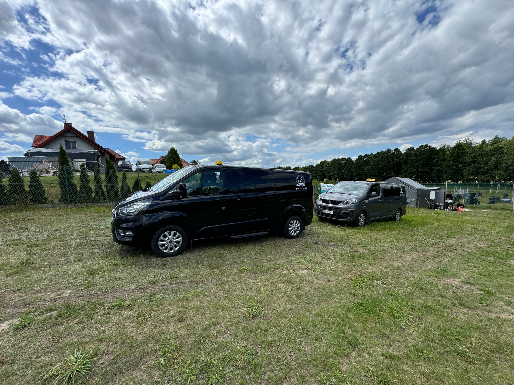 Dwa busy, czarny i szary, stoją na trawiastym terenie pod błękitnym niebem z chmurami; czarny bus z widocznym logo firmy na boku.