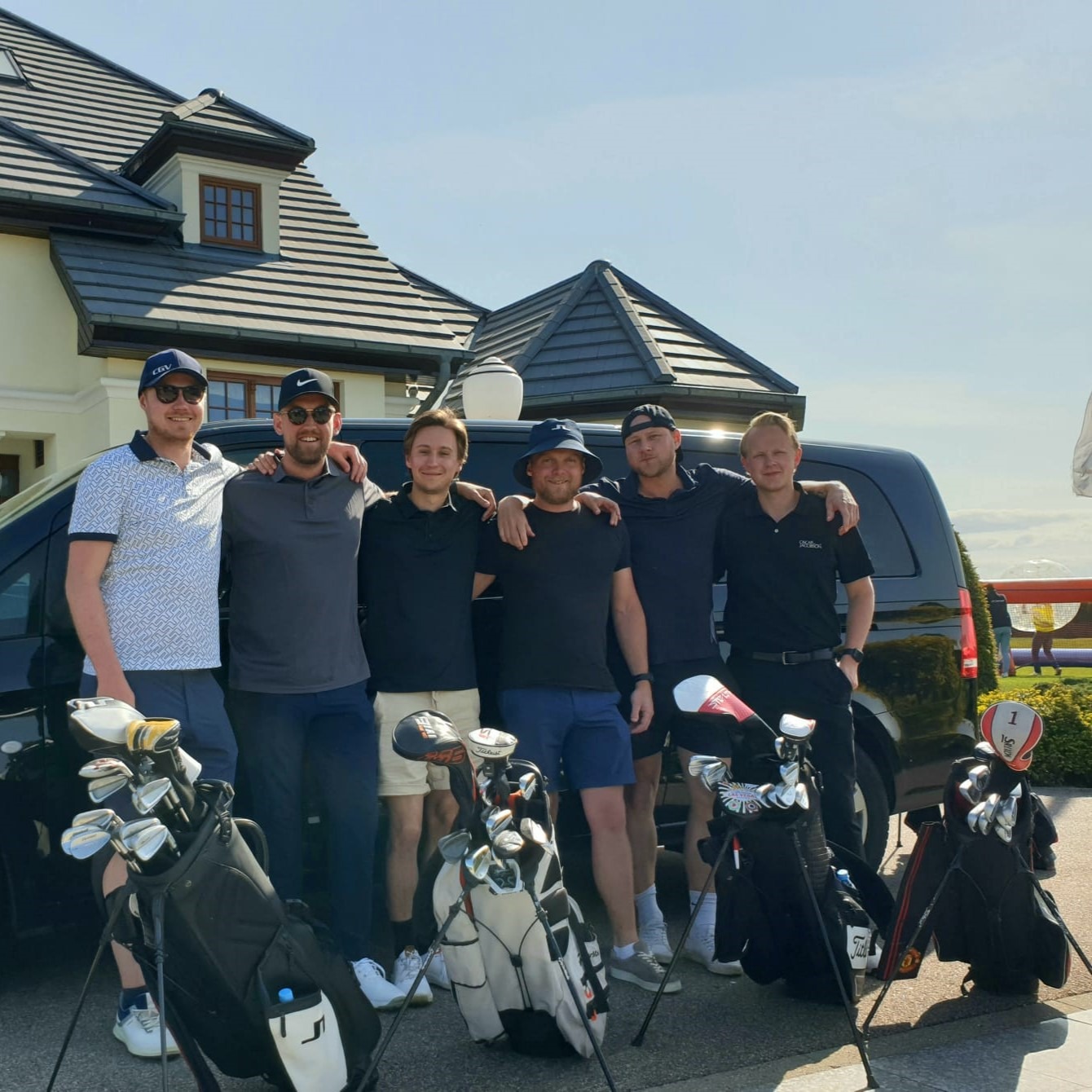 Grupa sześciu mężczyzn z torbami golfowymi pozuje przed czarnym busem w Gdańsku, prawdopodobnie po zakończonej grze w golfa.
