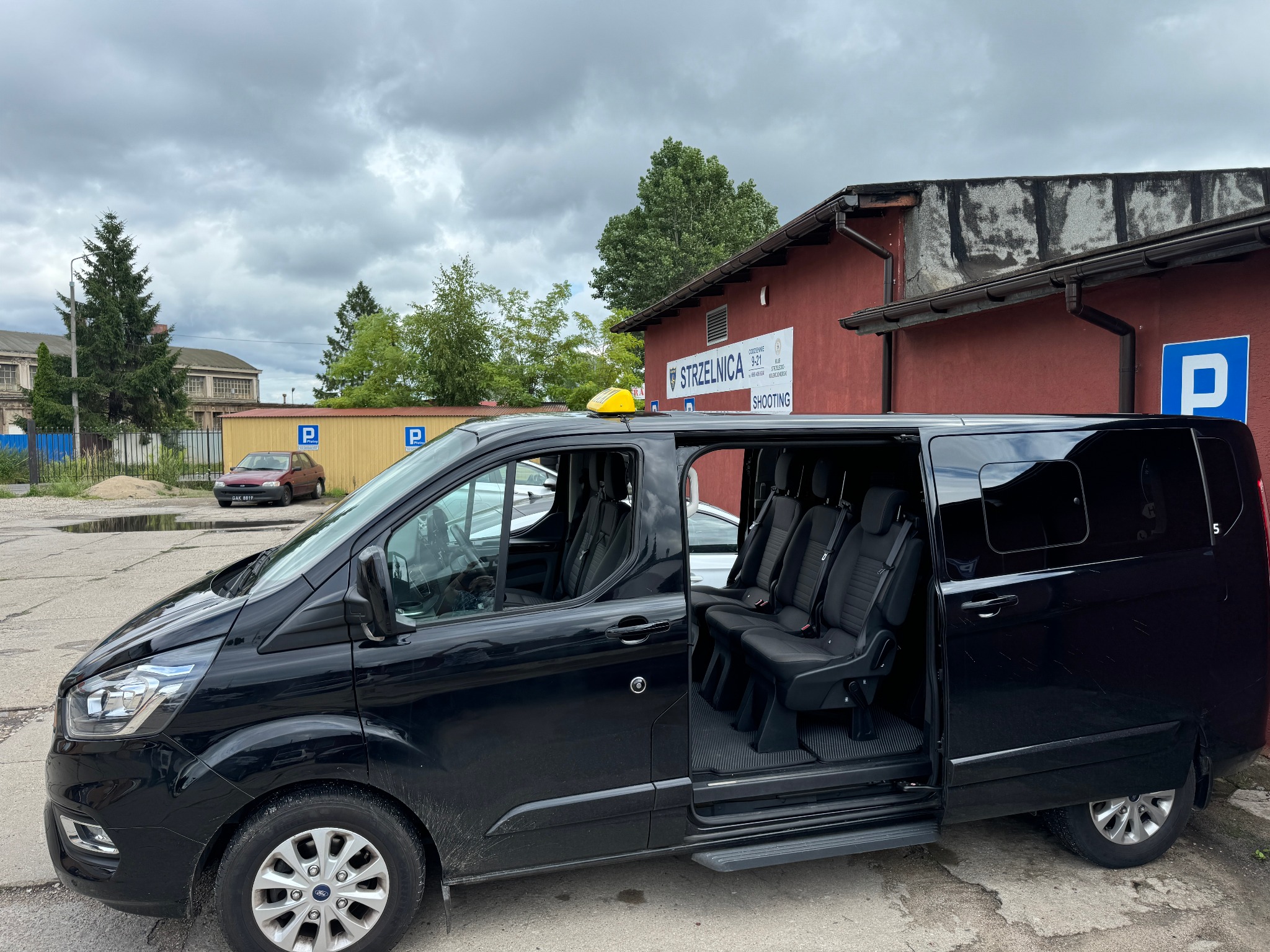 Czarny bus z otwartymi drzwiami przesuwnymi, widoczne wnętrze z fotelami pasażerskimi, żółta lampa taxi na dachu, budynek z napisem 'Strzelnica' w tle.