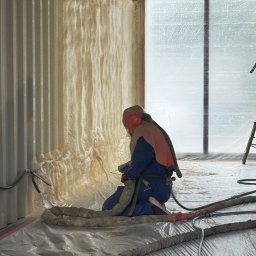 PurSolutions - Pracownik w kombinezonie ochronnym aplikuje piankę PUR na ścianę wewnątrz budynku. Widoczne węże, podłoga zabezpieczona folią. Ocieplanie pianką PUR.