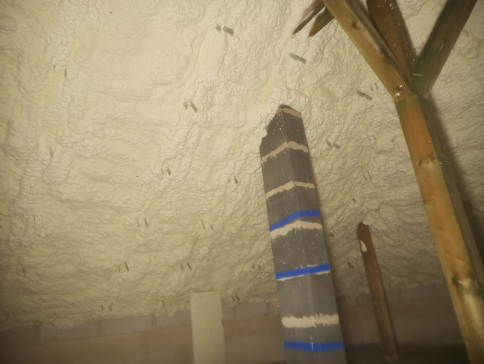 Poddasze po aplikacji pianki PUR, widoczne krokwie i element konstrukcyjny z taśmą malarską. Biała, nierówna powierzchnia izolacji.