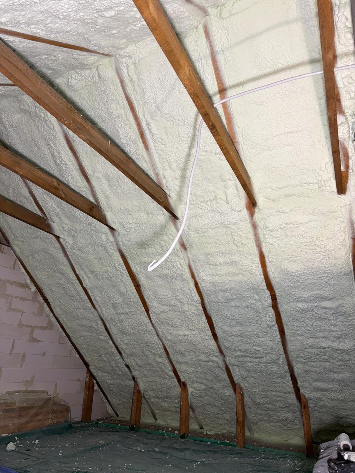 Poddasze po ociepleniu pianką PUR, widoczne drewniane krokwie i biała, spieniona izolacja. Kabel elektryczny zwisa z sufitu. Częściowo widoczna ściana z bloczków.
