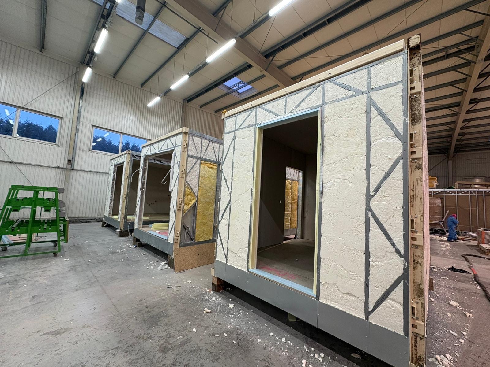 Modułowe ściany domu w hali produkcyjnej, z widoczną izolacją piankową i elementami konstrukcyjnymi. W tle okna i oświetlenie hali, częściowo wykończone moduły.
