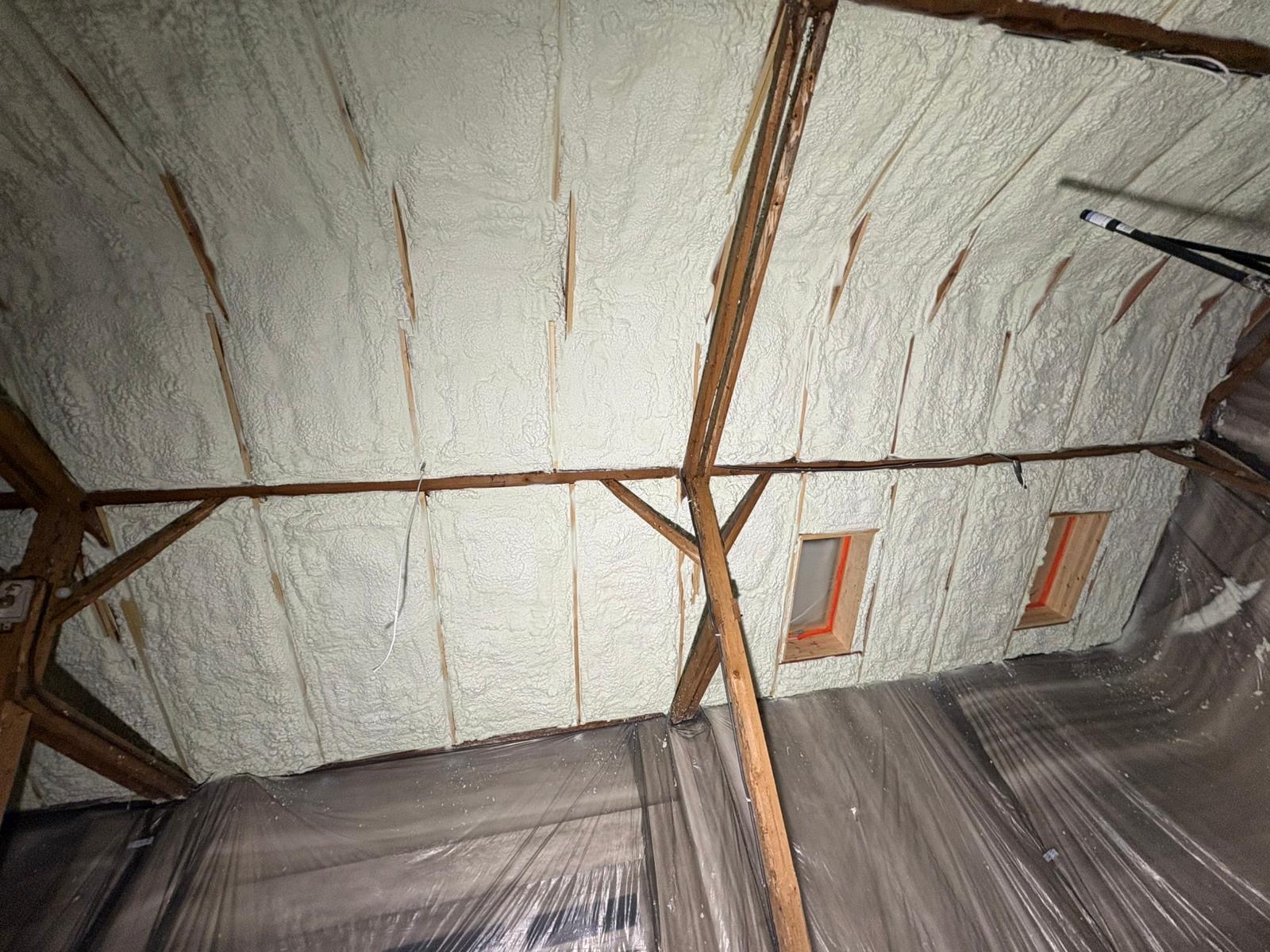 Poddasze z drewnianą konstrukcją i oknami połaciowymi, pokryte grubą warstwą jasnej pianki izolacyjnej. Widoczna folia ochronna na podłodze.