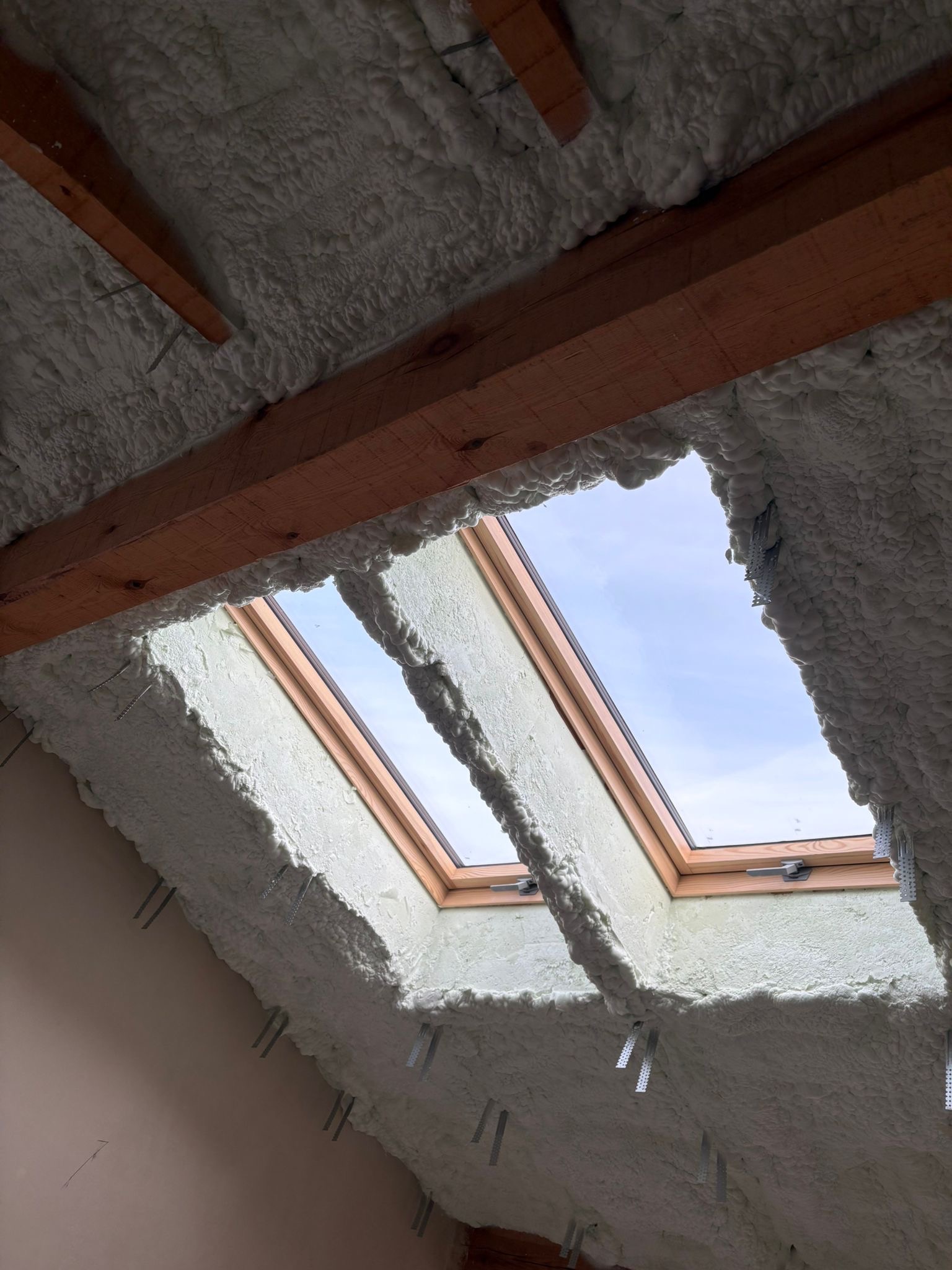 Poddasze z oknem dachowym po aplikacji pianki PUR, widoczne drewniane elementy konstrukcyjne i metalowe mocowania. Izolacja natryskowa, jasna piana.