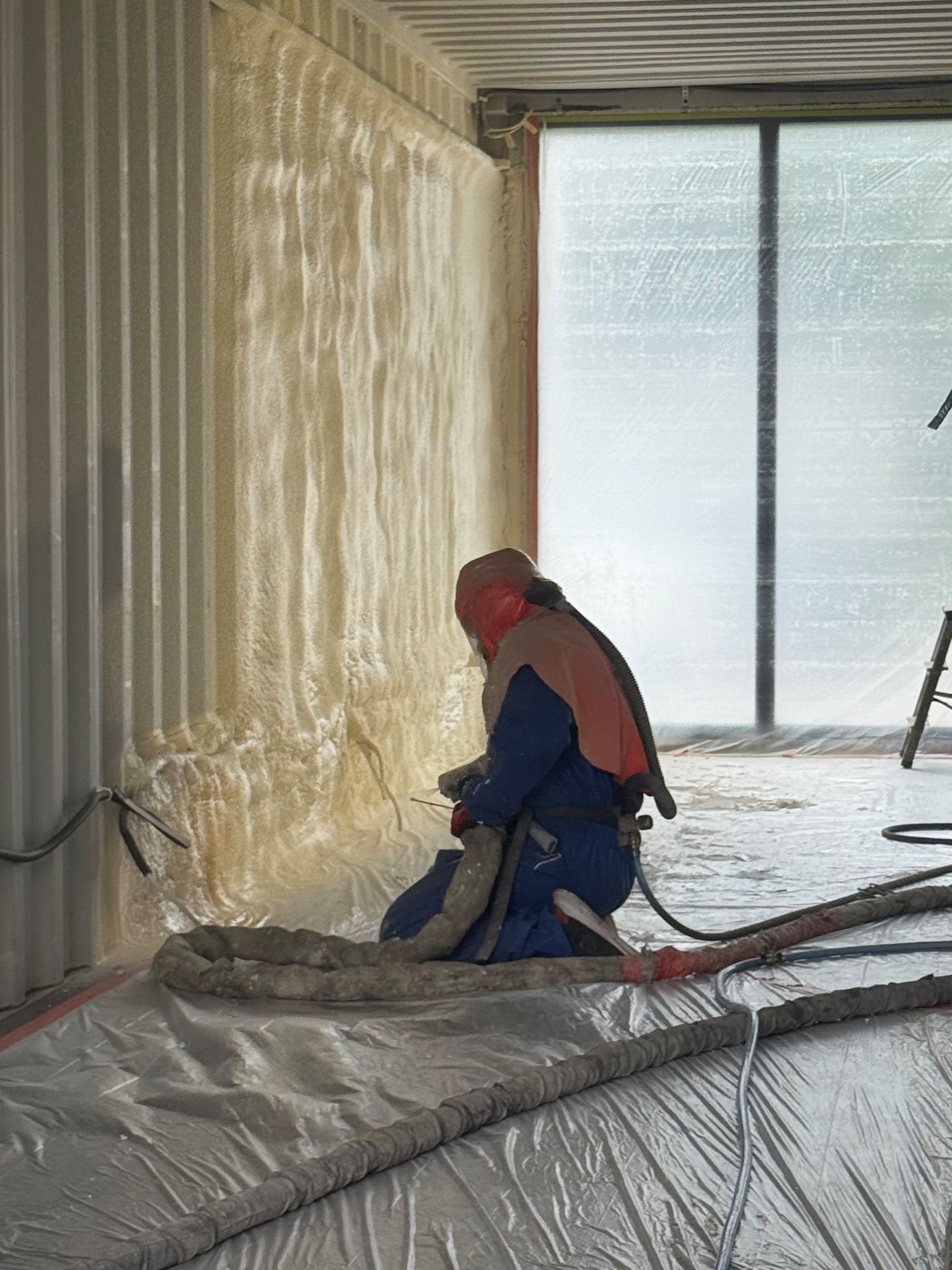 Pracownik w kombinezonie ochronnym aplikuje piankę PUR na ścianę wewnątrz budynku. Widoczne węże, podłoga zabezpieczona folią. Ocieplanie pianką PUR.