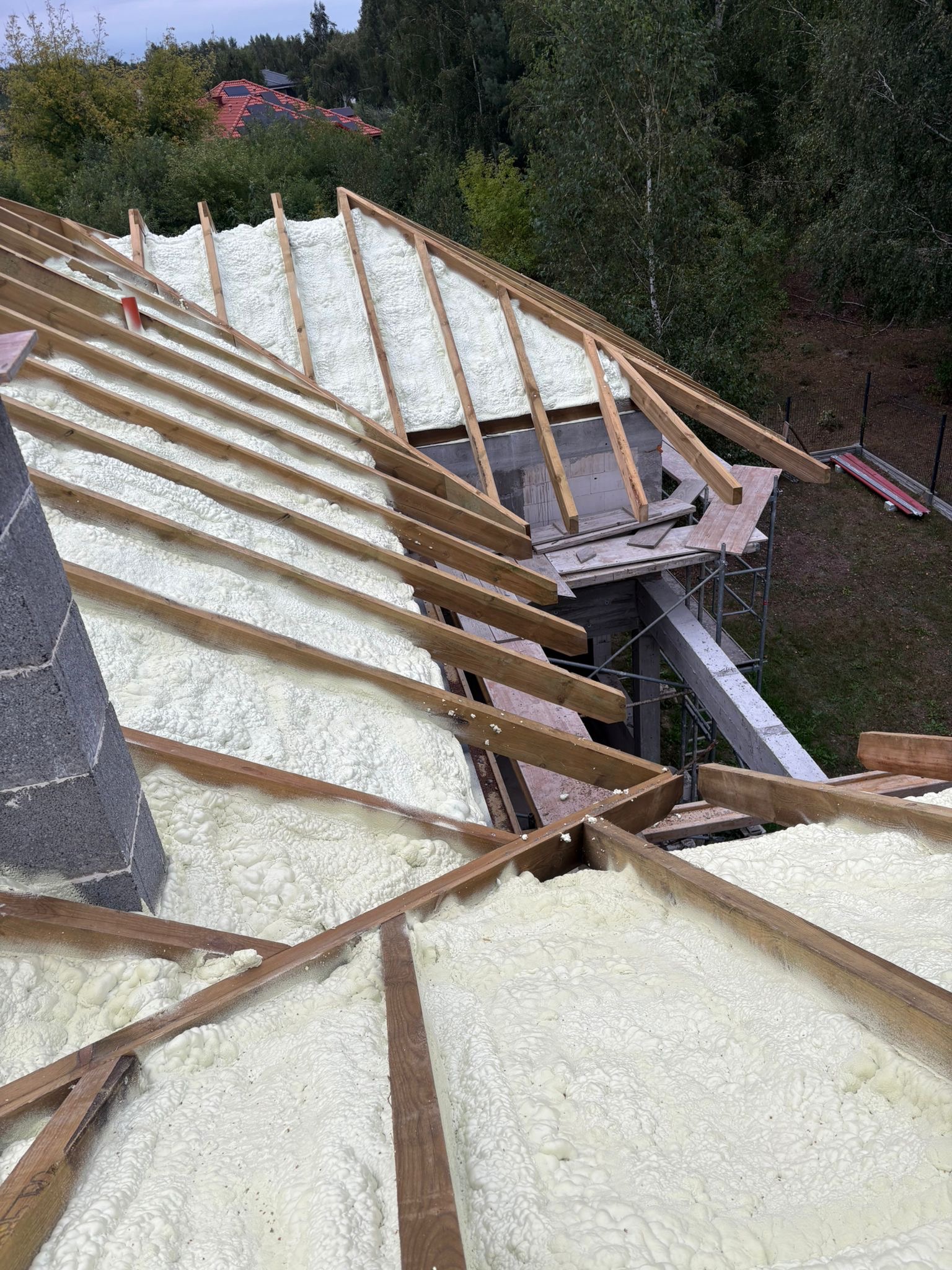Poddasze z drewnianą konstrukcją wypełnione białą pianką izolacyjną, widok z góry na ocieplony dach w trakcie prac budowlanych. Fragment komina z szarej cegły.