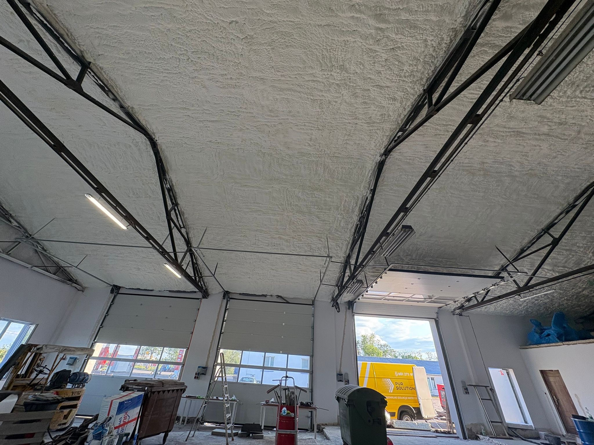 Poddasze hali przemysłowej po aplikacji pianki PUR, widoczne metalowe elementy konstrukcyjne i oświetlenie. W tle brama garażowa i fragment żółtego samochodu.
