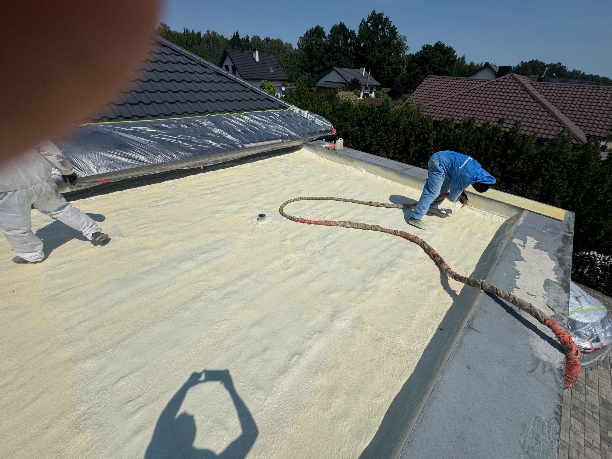 Aplikacja jasnej pianki izolacyjnej na dachu domu przez dwóch pracowników w kombinezonach ochronnych. Widoczne domy w tle i cień osoby robiącej zdjęcie.