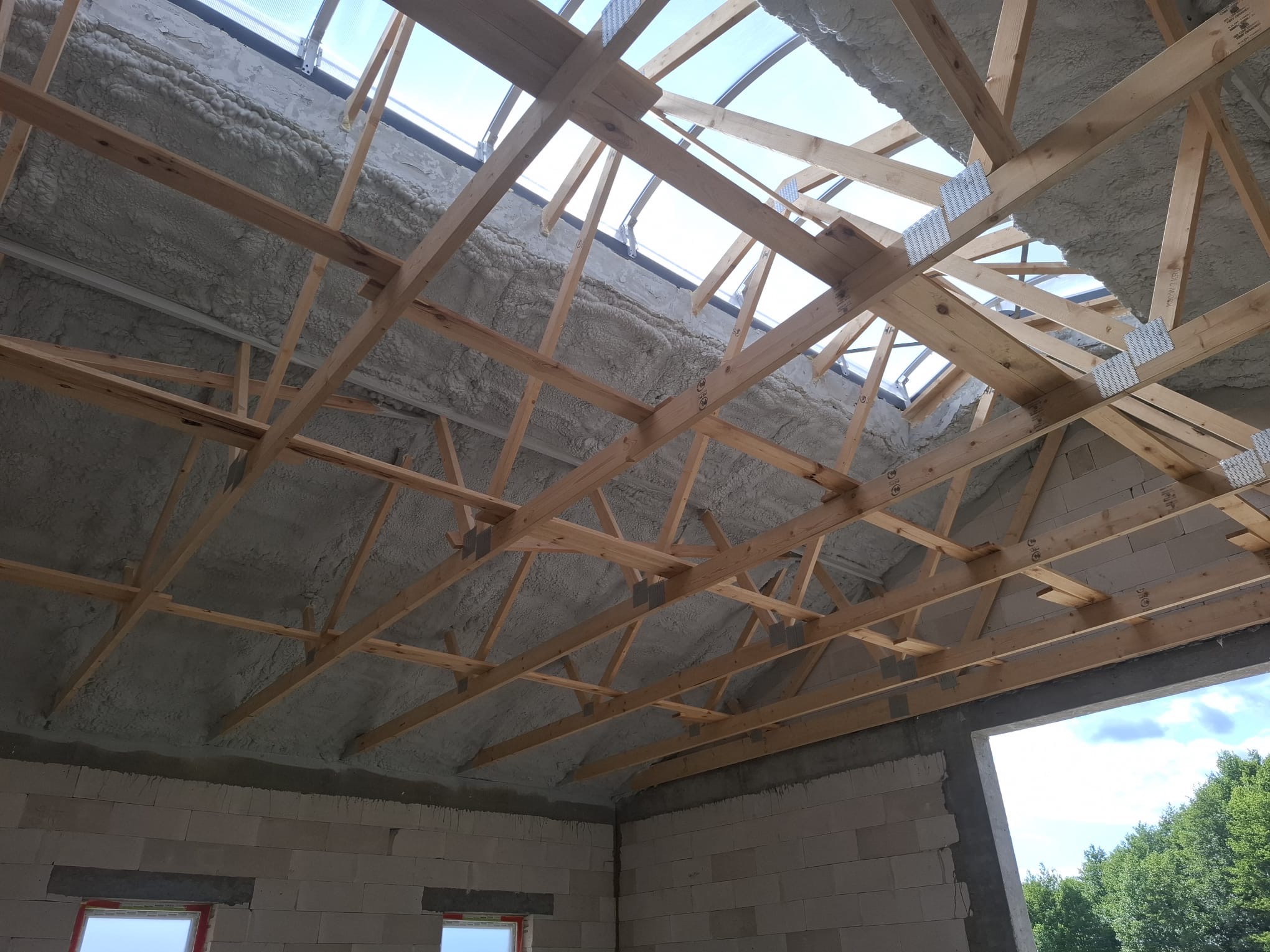 Poddasze z drewnianą konstrukcją dachu, ocieplone szarą pianką PUR, widoczne okno dachowe i fragment ściany z pustaków. Ocieplenie wykonane natryskowo.