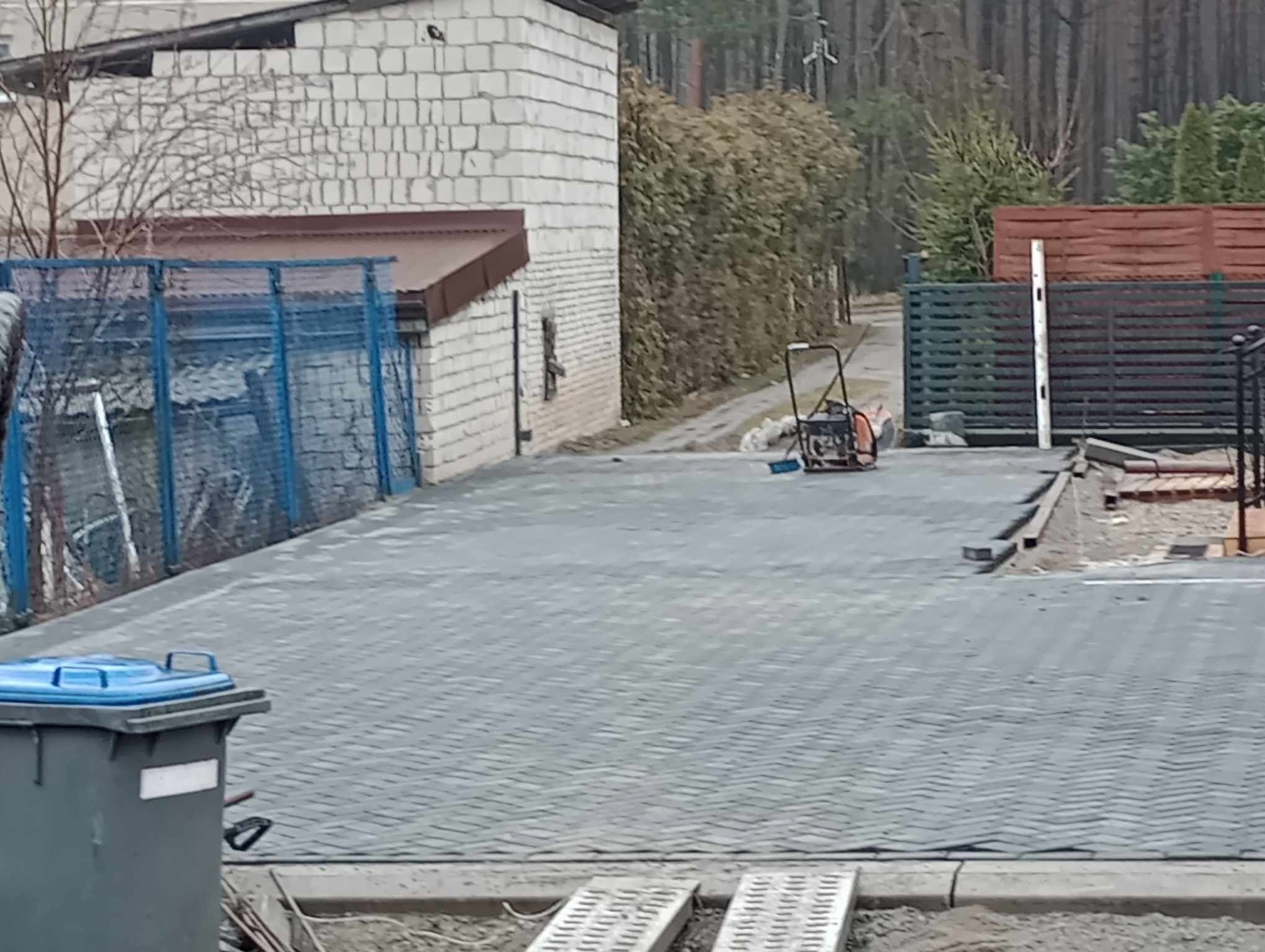 Nowo ułożona, szara kostka brukowa na podjeździe, widoczna zagęszczarka w tle, obok budynek z białej cegły i niebieskie ogrodzenie.