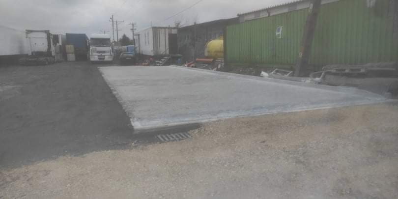 Wyrównany, szary, betonowy plac na terenie przemysłowym z zaparkowanymi ciężarówkami i kontenerami w tle.