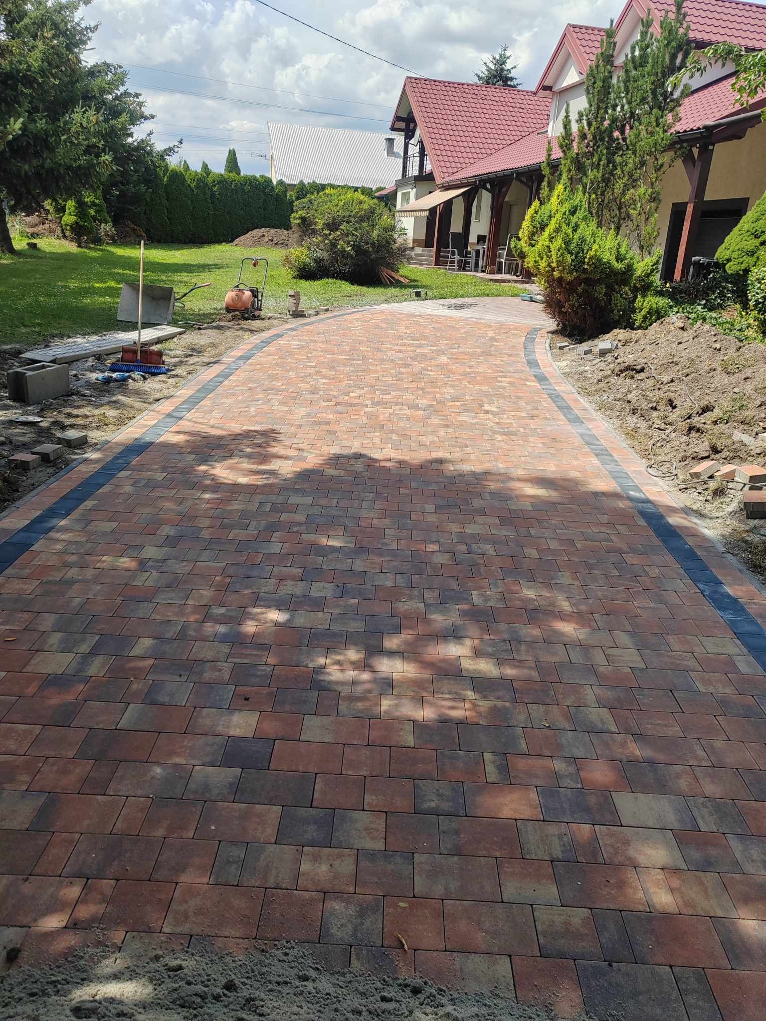 Zakrzywiony podjazd z kostki brukowej w odcieniach czerwieni i brązu, obrzeża z ciemnej kostki, widoczne narzędzia i maszyna do ubijania gruntu w tle, częściowo ułożona kostka po bokach.