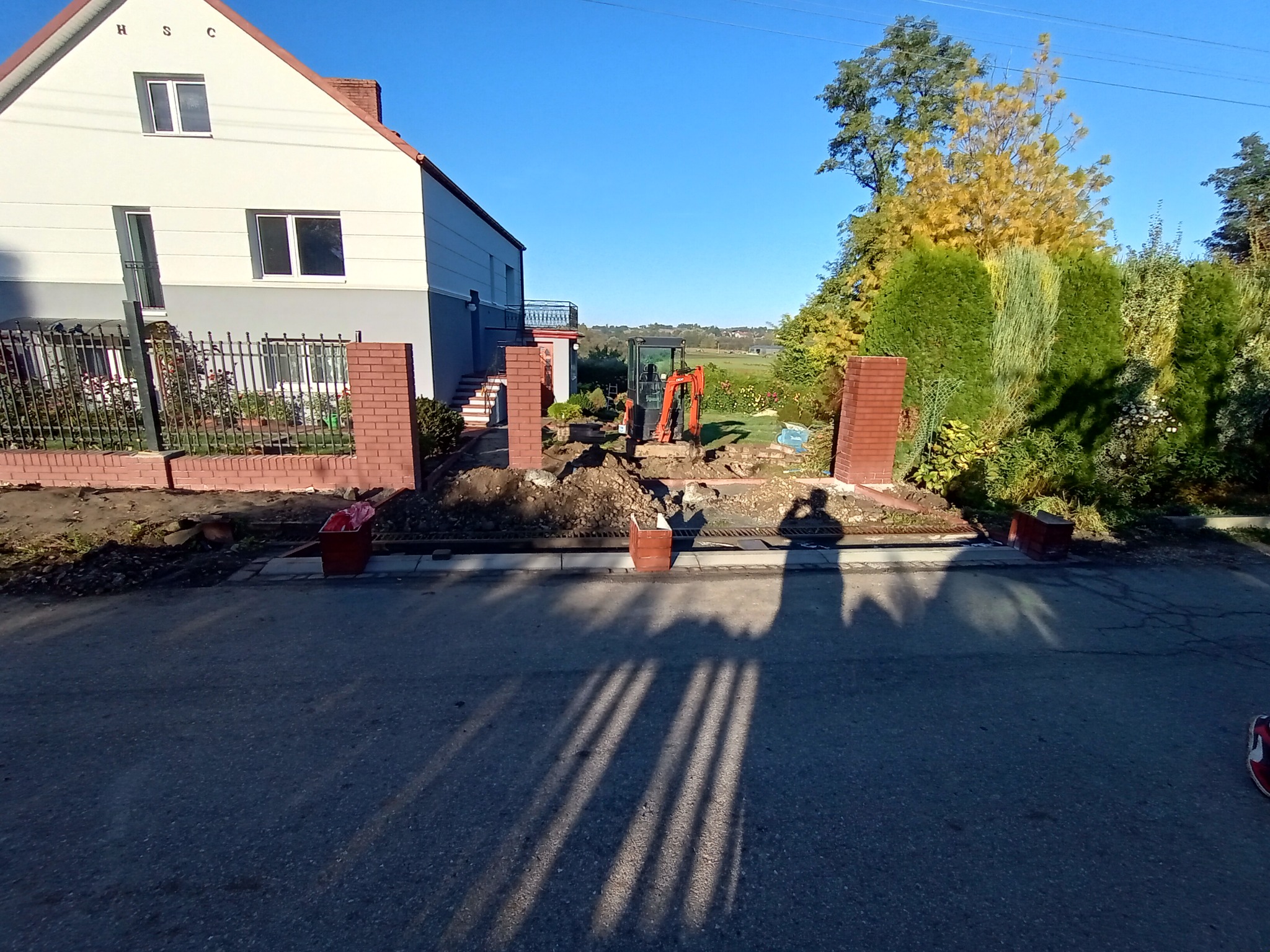 Mini koparka na działce przed domem, przygotowanie terenu pod budowę ogrodzenia z ceglanymi słupkami, widoczne wykopane fragmenty ziemi i cień osoby robiącej zdjęcie.