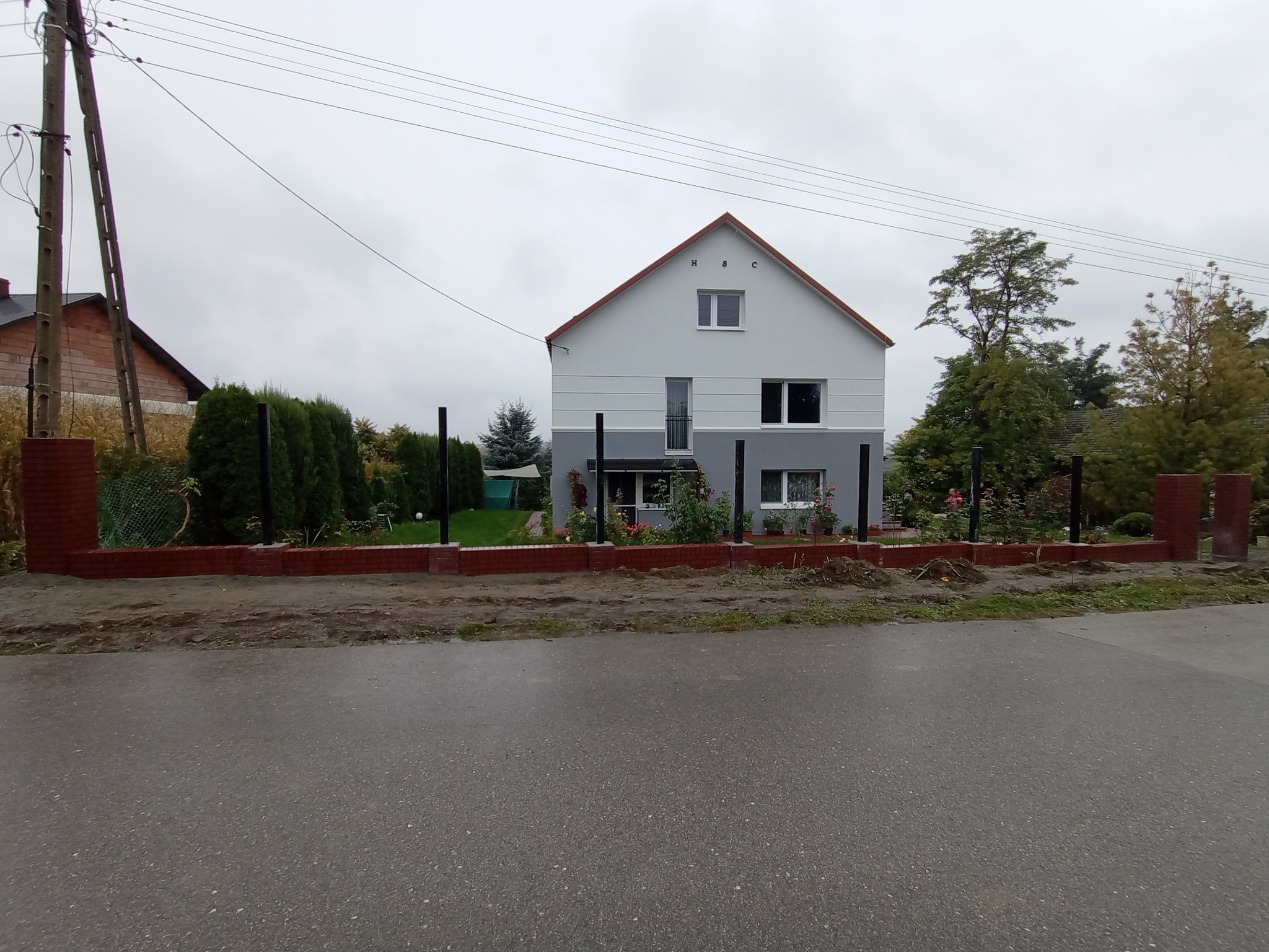 Nowoczesny dom z szarą elewacją i czerwoną ceglaną podmurówką oraz czarnym ogrodzeniem panelowym na tle pochmurnego nieba.