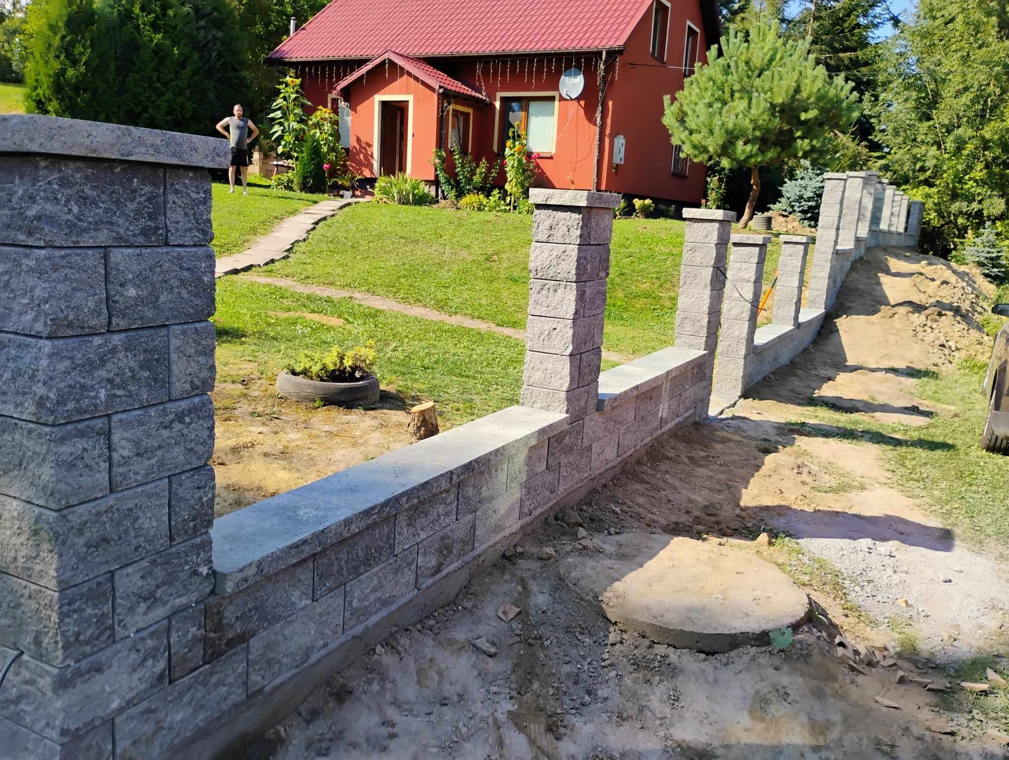 Wznoszenie muru oporowego z szarej kostki łupanej, widoczny fundament i słupki, w tle dom z czerwonym dachem i mężczyzna w ogrodzie.