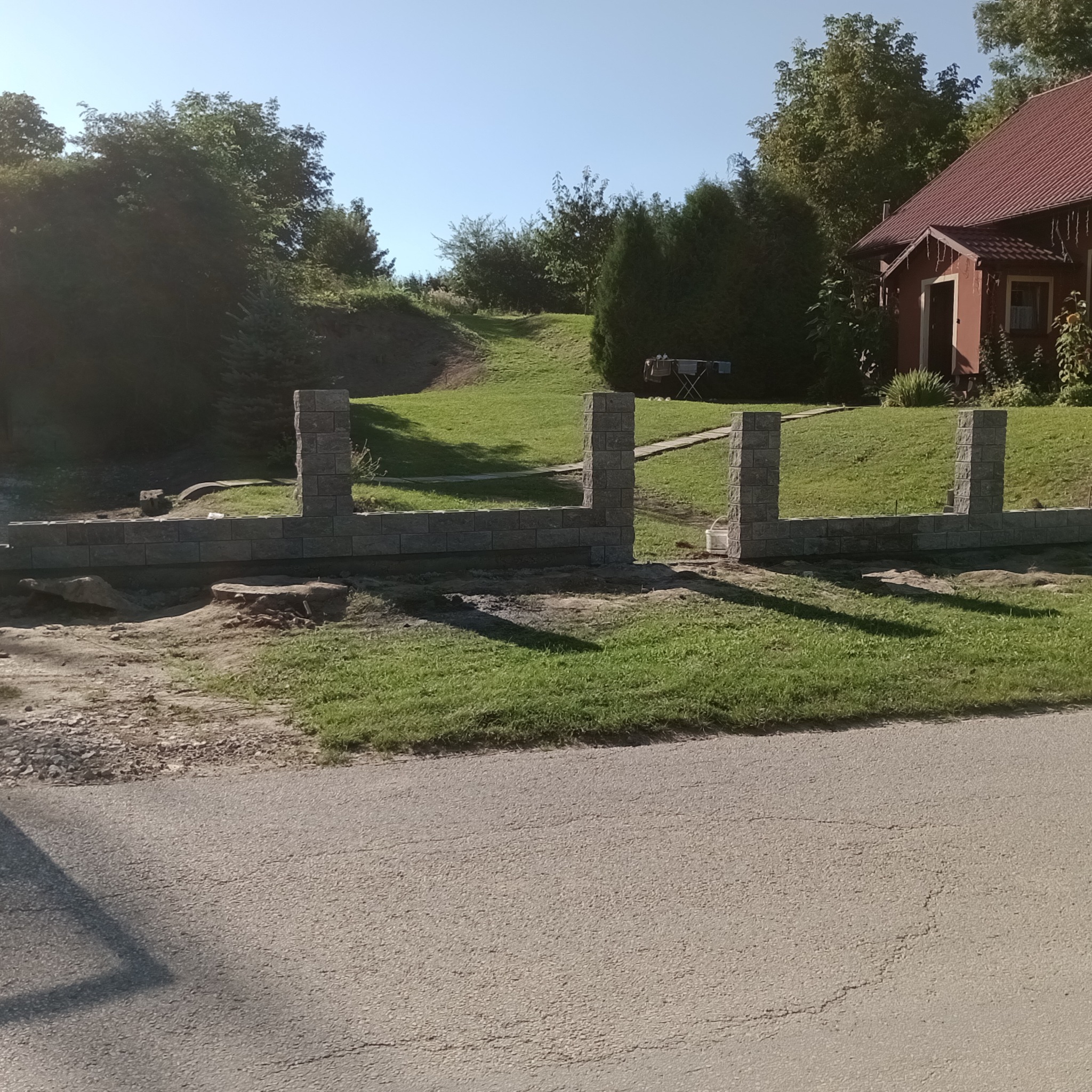 Wznoszenie murowanego ogrodzenia z szarej kostki betonowej, widoczne słupki i podmurówka w trakcie budowy, w tle trawnik i dom z czerwonym dachem.
