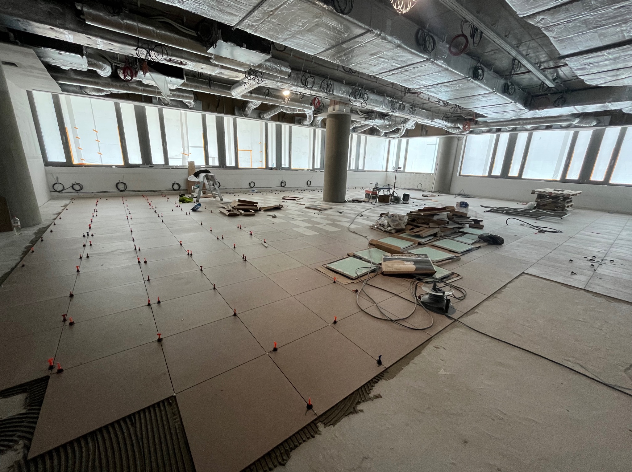 Układanie brązowych płytek podłogowych w dużym, jasnym pomieszczeniu z widocznymi elementami instalacji pod sufitem i dużymi oknami, z użyciem pomarańczowych klinów poziomujących.