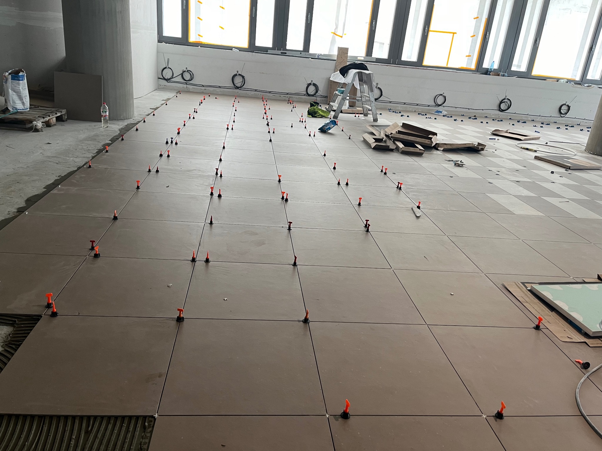 Układanie brązowych płytek podłogowych w dużym pomieszczeniu z widocznymi krzyżykami poziomującymi, pozostałościami kleju i częściowo ułożonymi płytkami, przy oknach drabina i narzędzia.