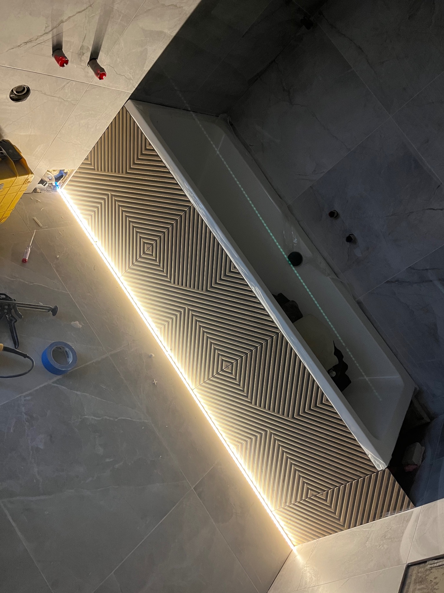 Wanna zabudowana płytkami z geometrycznym wzorem podświetlonym listwą LED, widoczne narzędzia i akcesoria montażowe na podłodze z płytek.