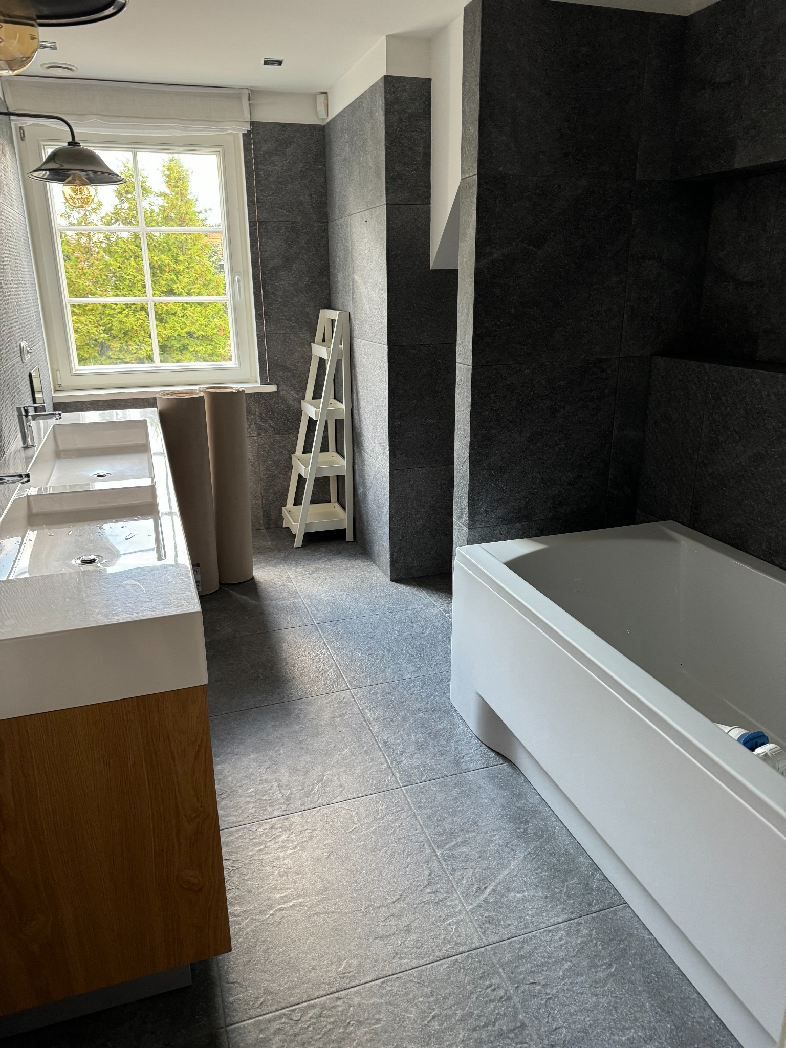 Nowoczesna łazienka w szarościach z białą wanną, umywalką z szafką w kolorze drewna i drabinką dekoracyjną; widoczne dwie rolki materiału budowlanego przy oknie.