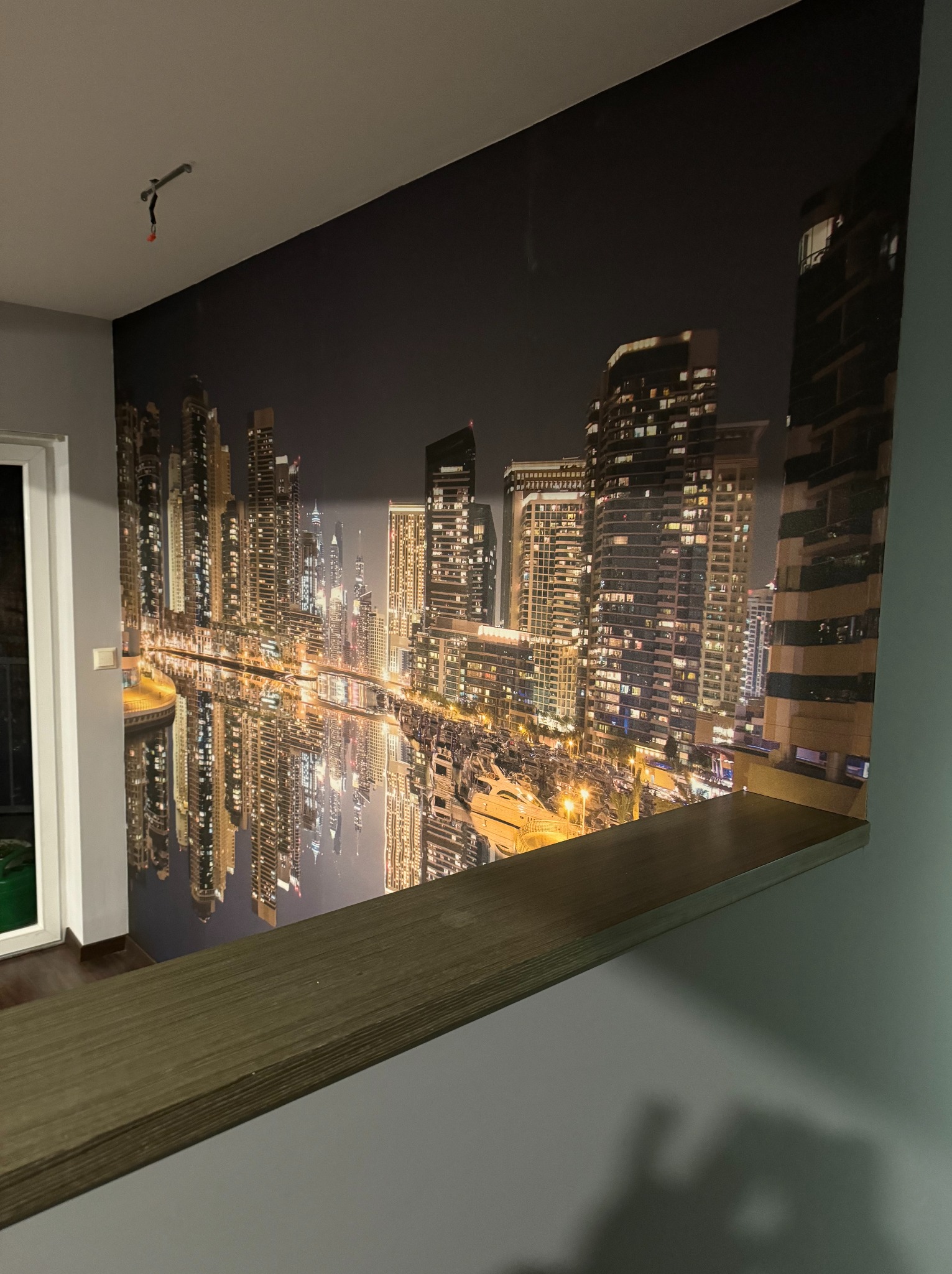 Fototapeta na ścianie przedstawiająca nocną panoramę Dubaju z odbiciem w wodzie, widoczna półka z ciemnego drewna na tle ściany.