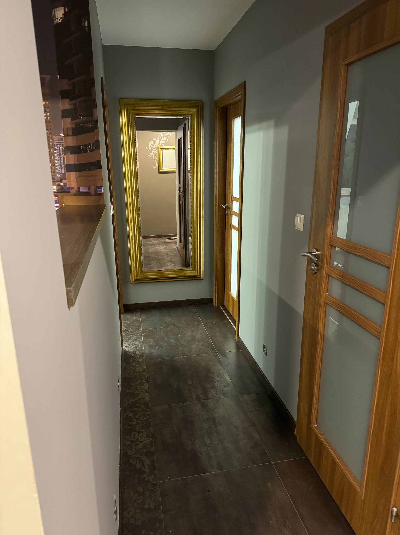 Wnętrze korytarza z ciemną podłogą, szarymi ścianami i drewnianymi drzwiami z przeszkleniami; w głębi widoczne lustro w złotej ramie.