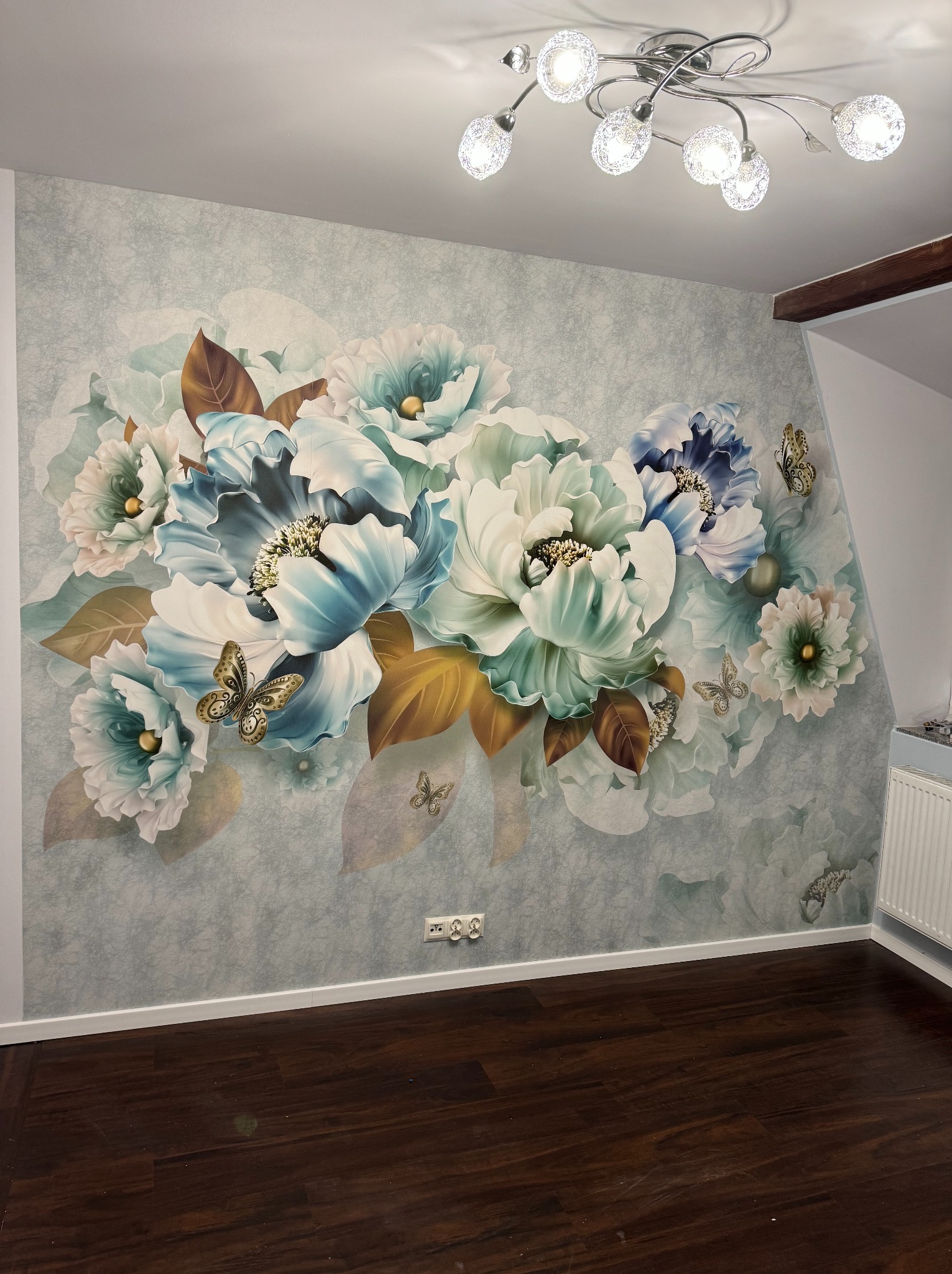 Ściana w pokoju z fototapetą przedstawiającą duże, stylizowane kwiaty w odcieniach błękitu i bieli z dodatkiem złotych liści i motyli, podłoga z ciemnego drewna, nowoczesna lampa sufitowa z wieloma...