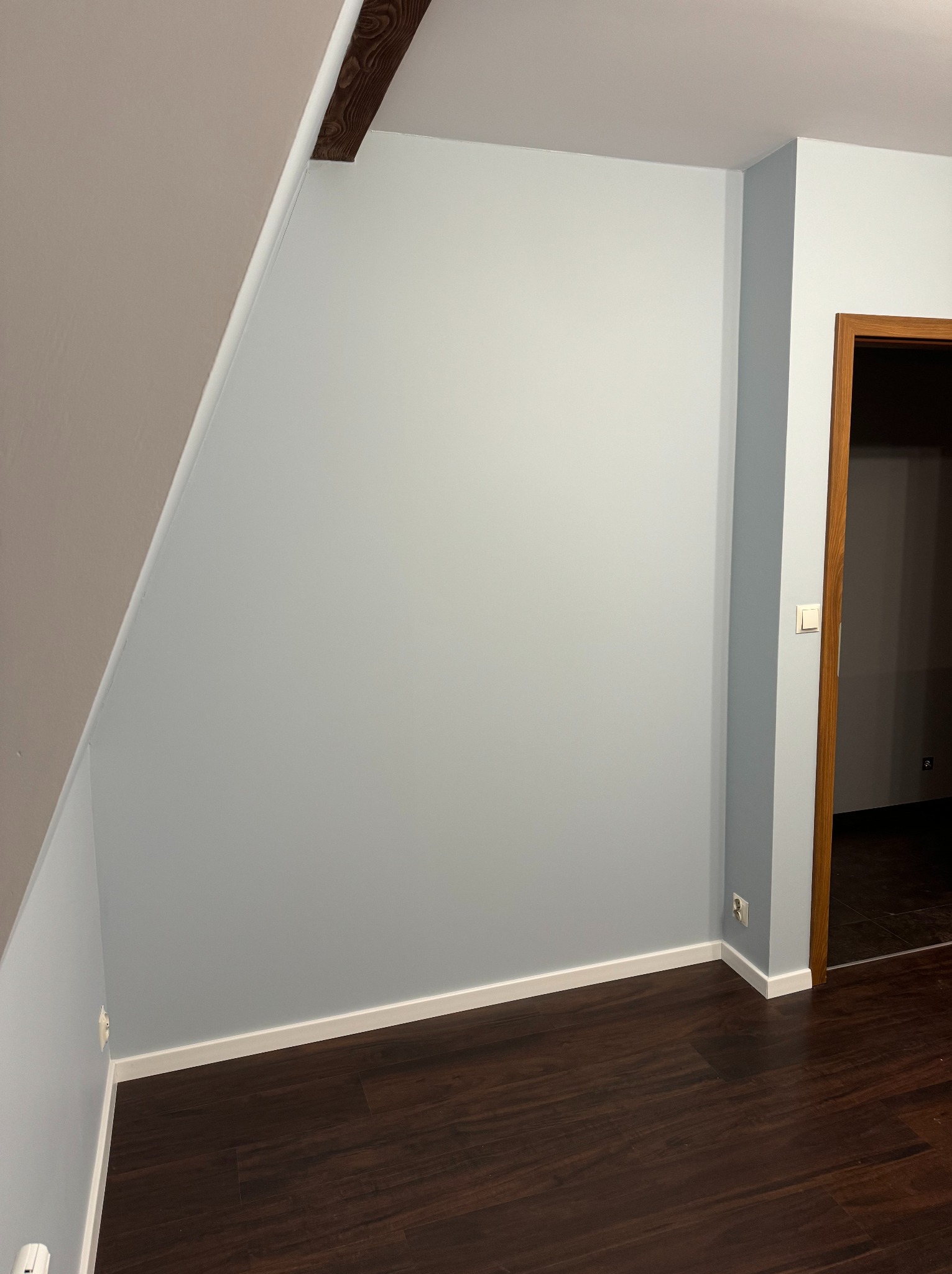 Świeżo pomalowane na szaro ściany w pokoju we Wrocławiu, z ciemnobrązową podłogą imitującą drewno i fragmentem drewnianej belki stropowej przy skośnym suficie, widok na otwarte drzwi do sąsiedniego...