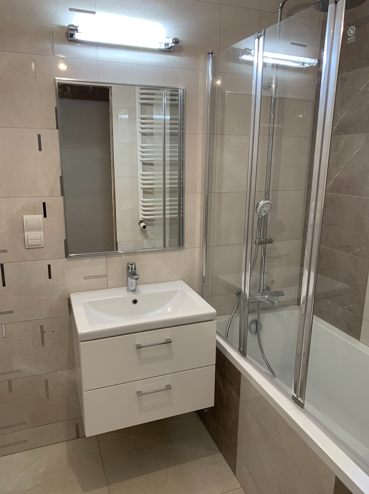 Nowoczesna łazienka z beżowymi i brązowymi płytkami, białą szafką pod umywalką z szufladami i kabiną prysznicową z elementami chromowanymi.