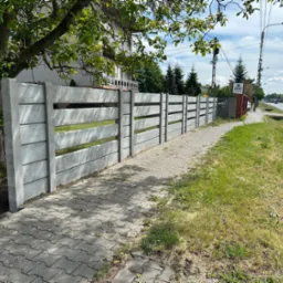 Nowoczesny, prefabrykowany, betonowy płot panelowy z poziomymi szczelinami, oddzielający posesję od chodnika i ulicy, widok z boku, w tle dom i drzewa.