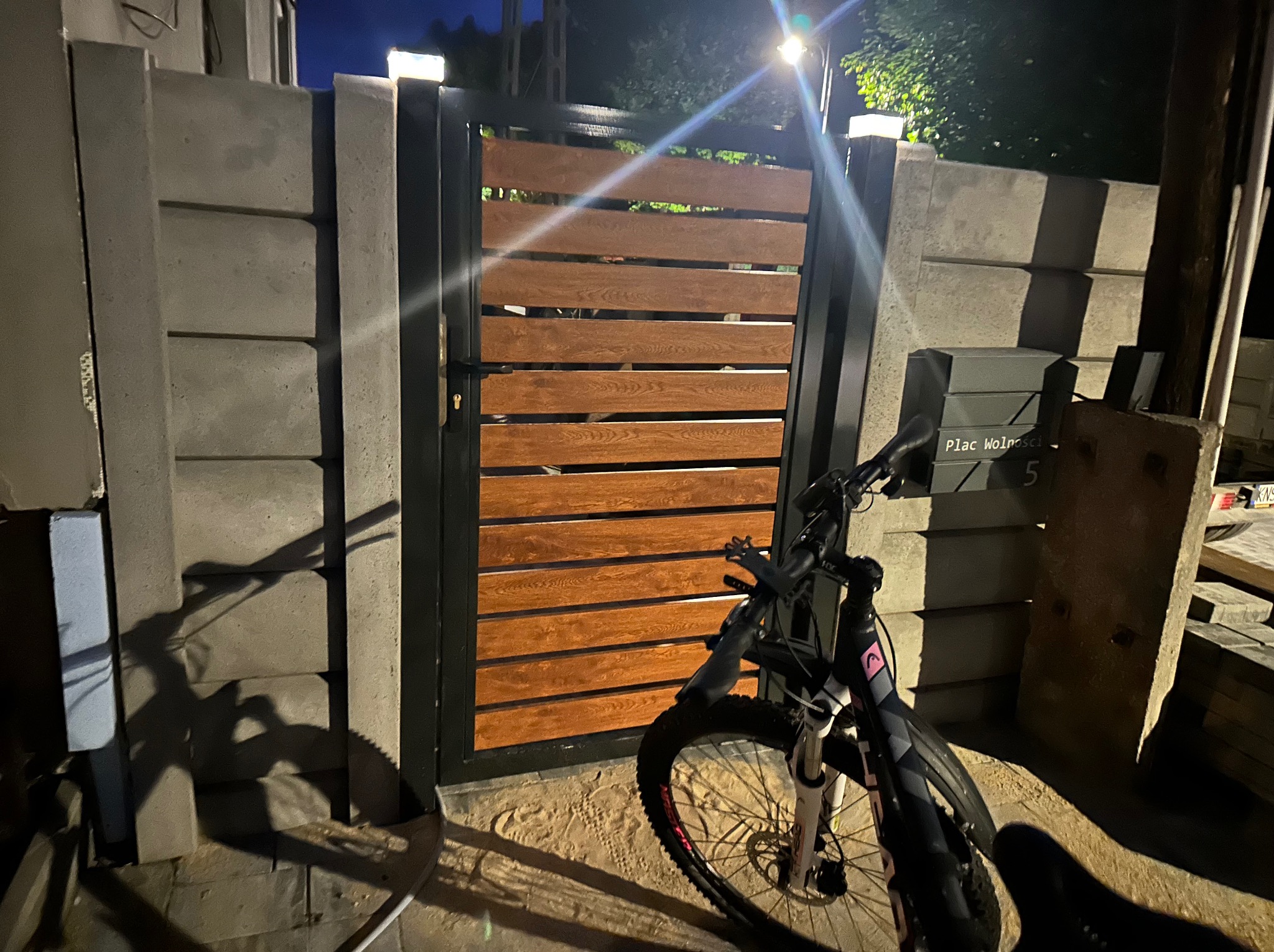Nowoczesna brama ogrodzeniowa z poziomych drewnianych paneli w czarnej ramie, betonowe słupki z oświetleniem, rower górski zaparkowany przed bramą, widoczny adres na skrzynce pocztowej.