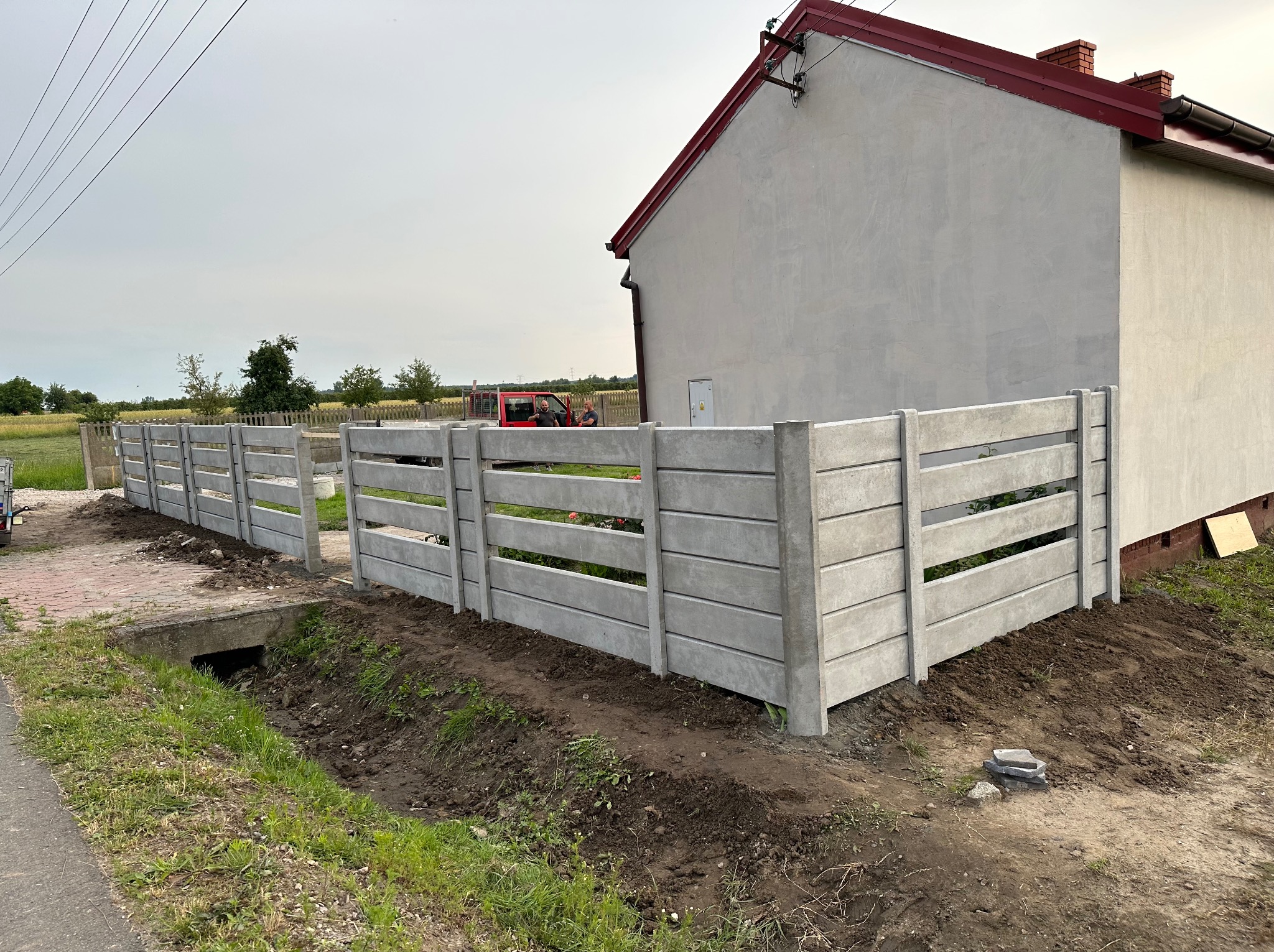 Nowoczesne ogrodzenie panelowe z prefabrykatów betonowych, widoczne z zewnątrz posesji, z fragmentem budynku w tle.