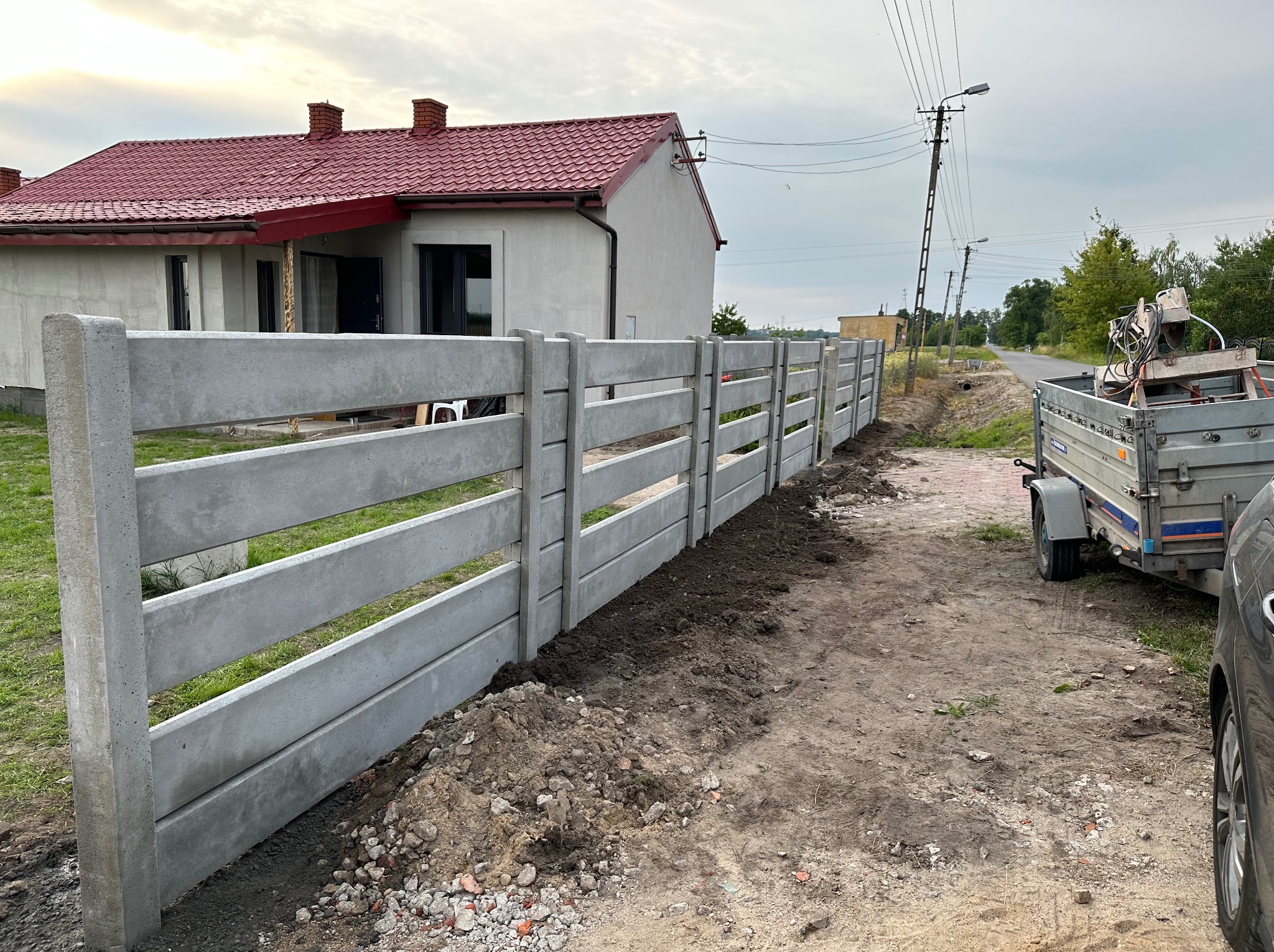 Nowo postawiony, szary, betonowy płot panelowy z widocznym domem z czerwoną dachówką w tle i przyczepką z narzędziami po prawej stronie.