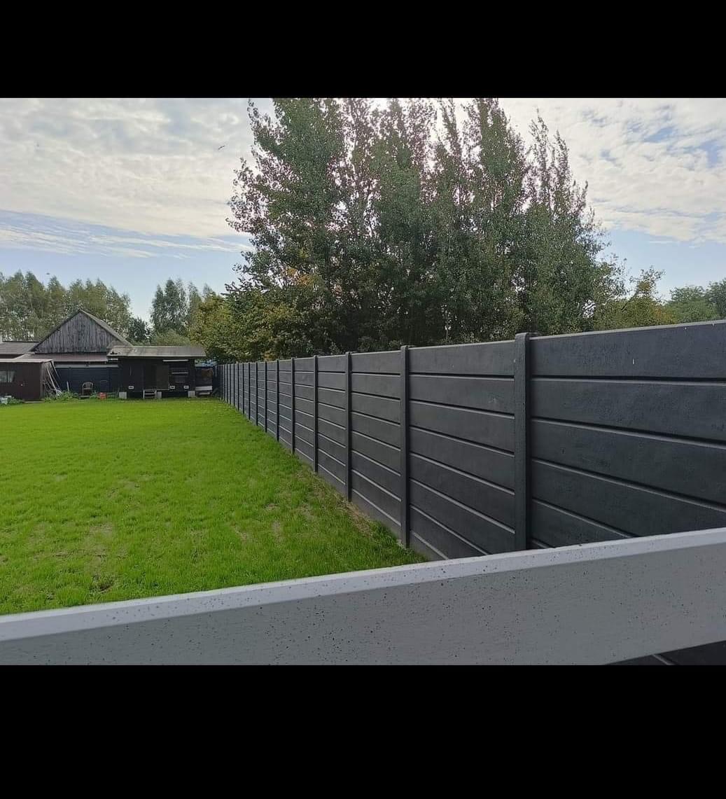 Nowoczesne, poziome, szare ogrodzenie panelowe z betonu architektonicznego, oddzielające zielony trawnik od zabudowań gospodarczych i drzew w tle, widoczne z perspektywy dolnej.