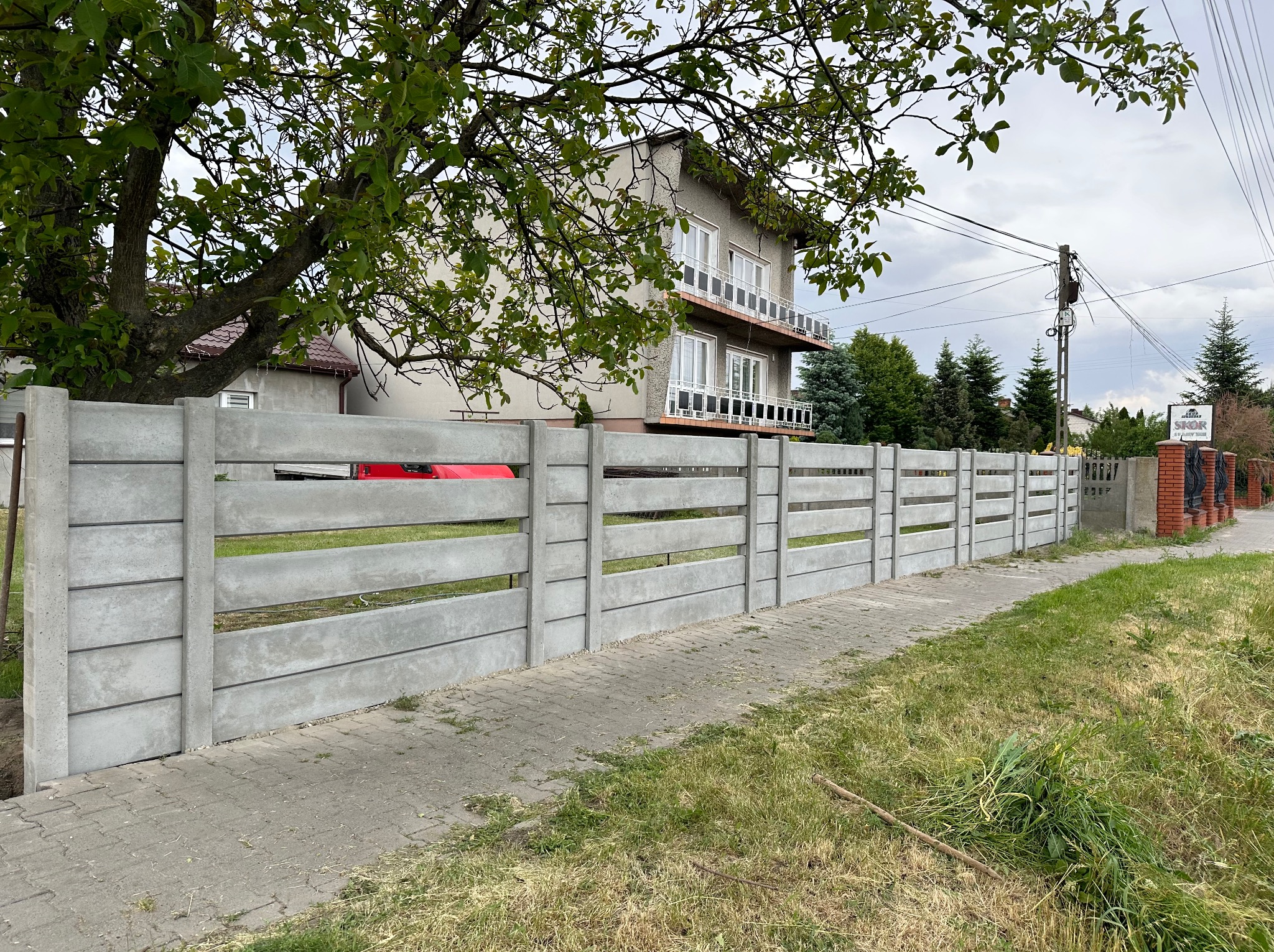 Nowoczesne, betonowe ogrodzenie panelowe z poziomymi przęsłami, widoczne z poziomu chodnika, z fragmentem czerwonego auta w tle.