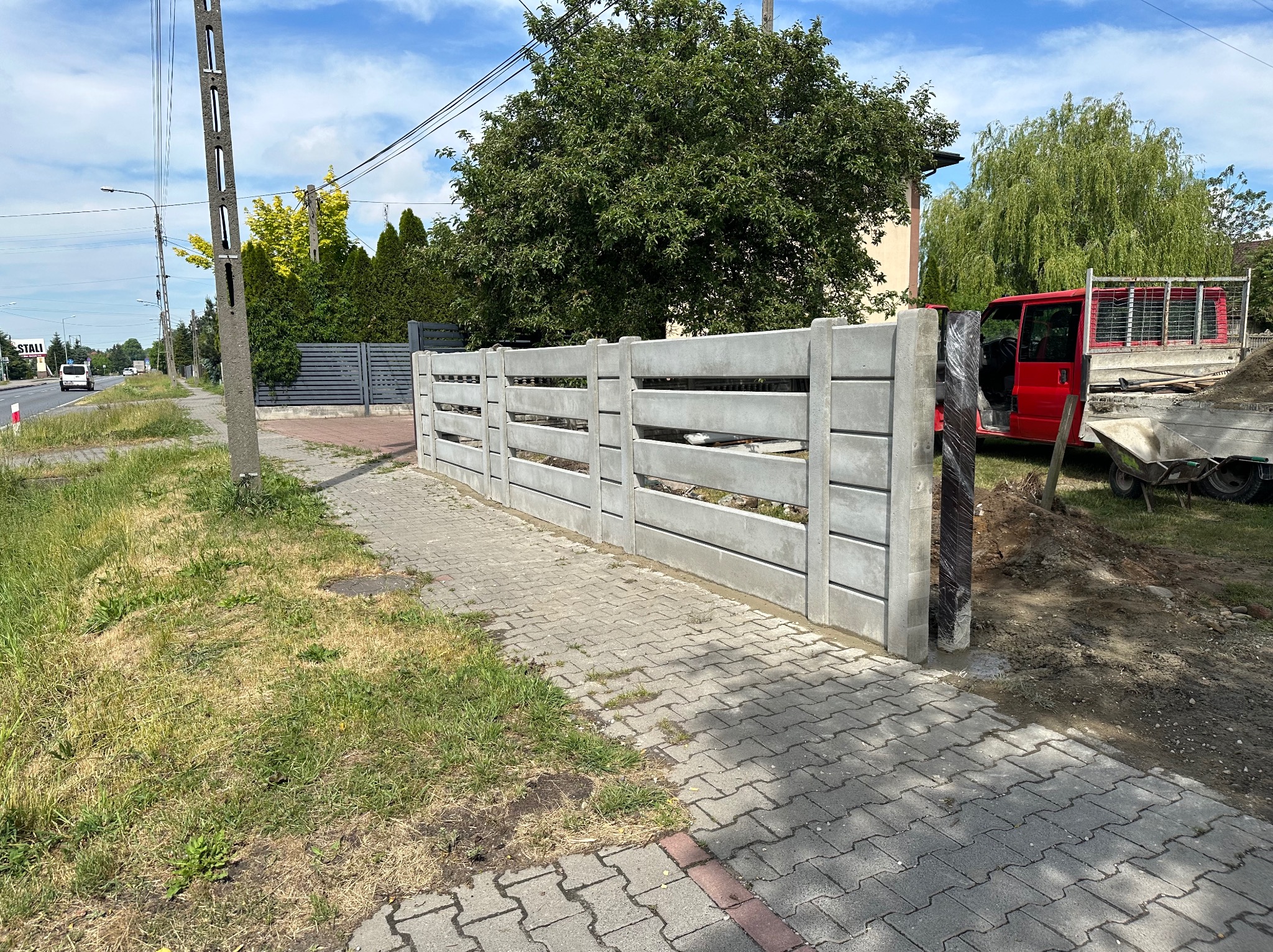 Nowoczesne, betonowe ogrodzenie panelowe w trakcie montażu, widoczny czerwony samochód dostawczy i materiały budowlane, trawnik i chodnik z kostki brukowej w tle.