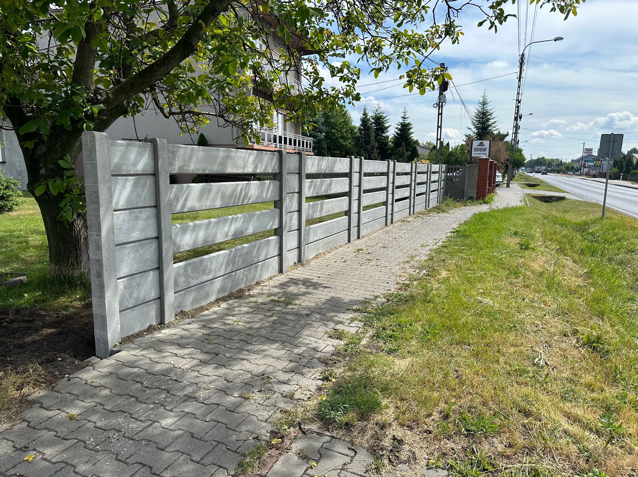 Nowoczesny, prefabrykowany, betonowy płot panelowy z poziomymi szczelinami, oddzielający posesję od chodnika i ulicy, widok z boku, w tle dom i drzewa.