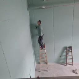 Pracownik na drabinie montuje płyty karton gips na ścianie, widoczne ślady po wkrętach, druga drabina oparta o ścianę w tle.