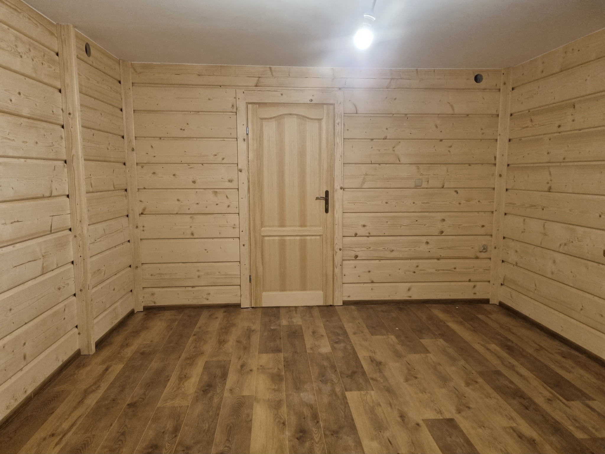 Wnętrze wykończone jasnym drewnem, z widoczną drewnianą podłogą, ścianami z bali i prostymi drewnianymi drzwiami z czarną klamką, oświetlone centralną lampą sufitową.