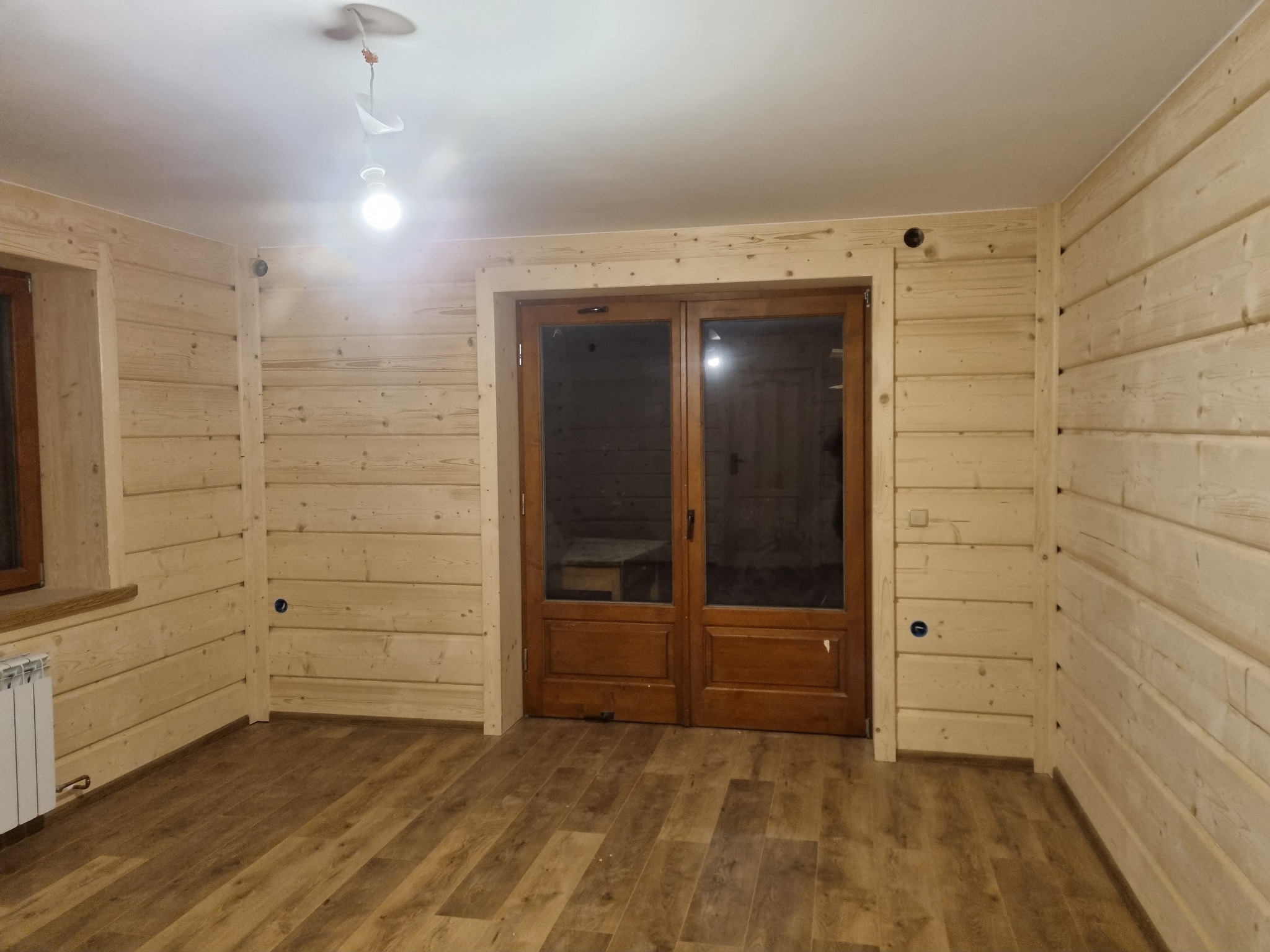 Wnętrze wykończone boazerią z jasnego drewna, z brązowymi, dwuskrzydłowymi drzwiami i panelami podłogowymi imitującymi drewno. Na suficie widoczne niedokończone podłączenie oświetlenia.