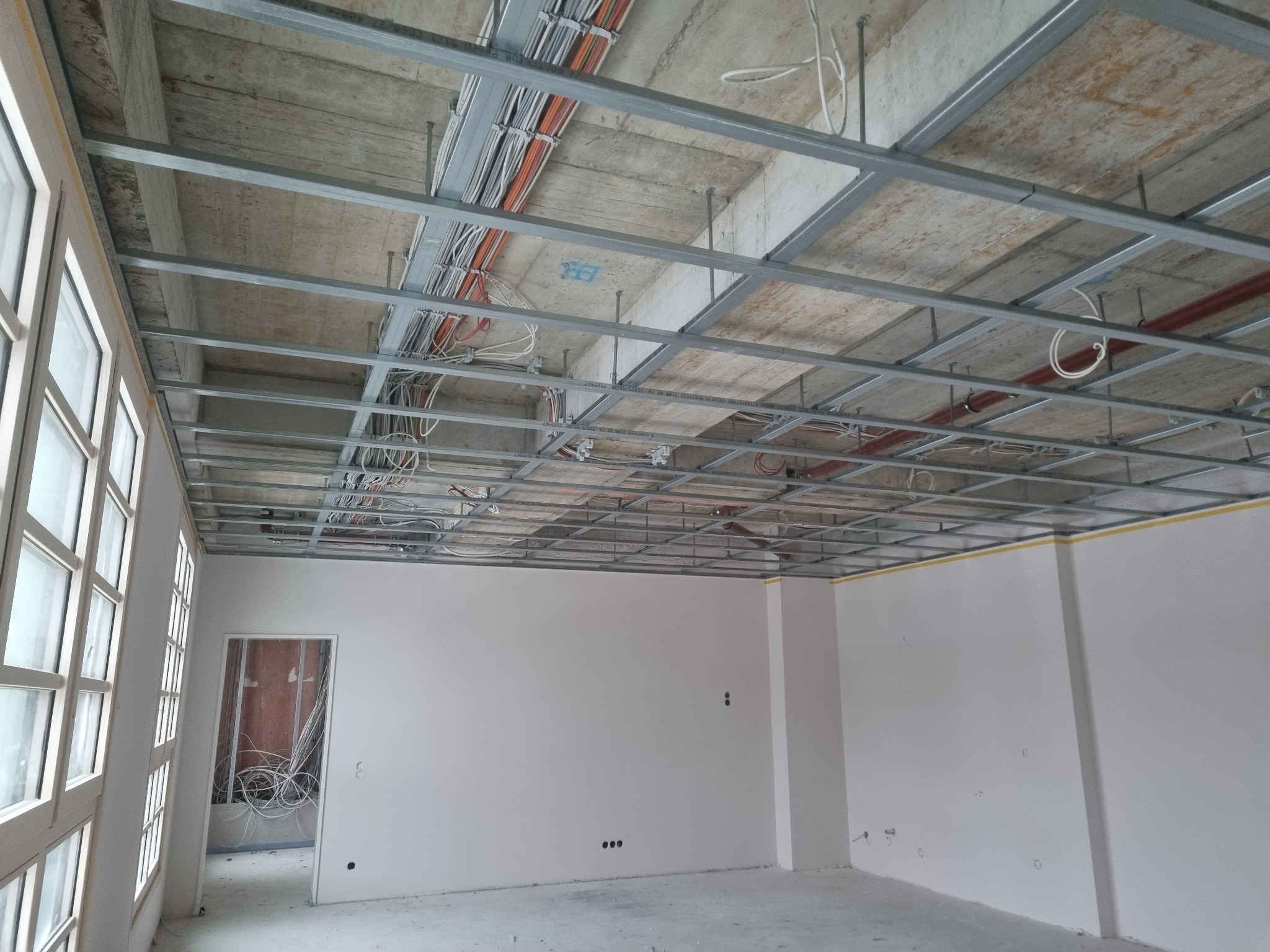 Stelaż sufitu podwieszanego z widocznymi przewodami elektrycznymi w surowym wnętrzu z oknami, przygotowany do montażu płyt kartonowo-gipsowych.