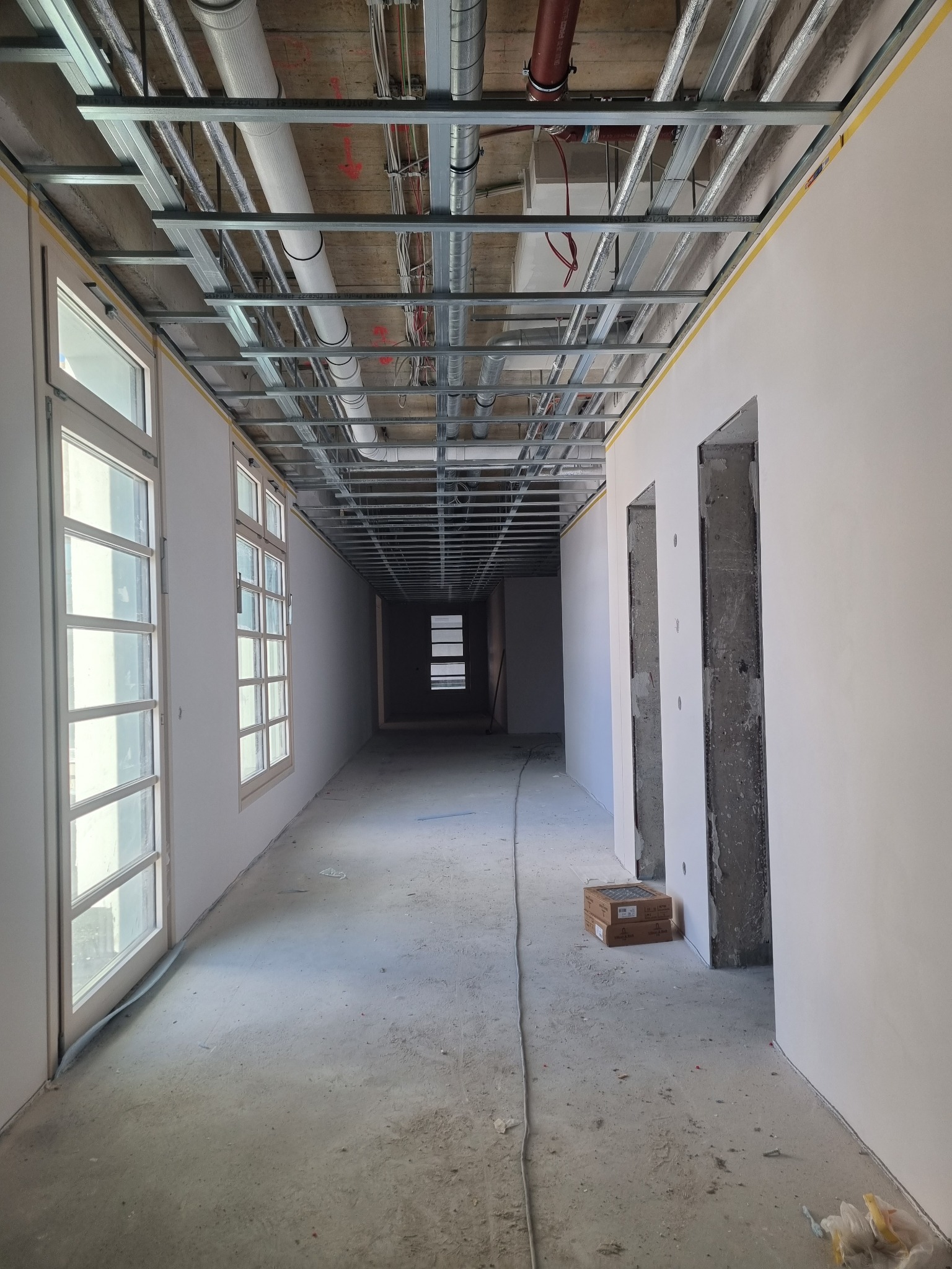 Korytarz w trakcie remontu: widoczne okna, surowy beton na podłodze, metalowa konstrukcja sufitu podwieszanego, instalacje pod sufitem oraz przygotowane otwory drzwiowe w ścianach.
