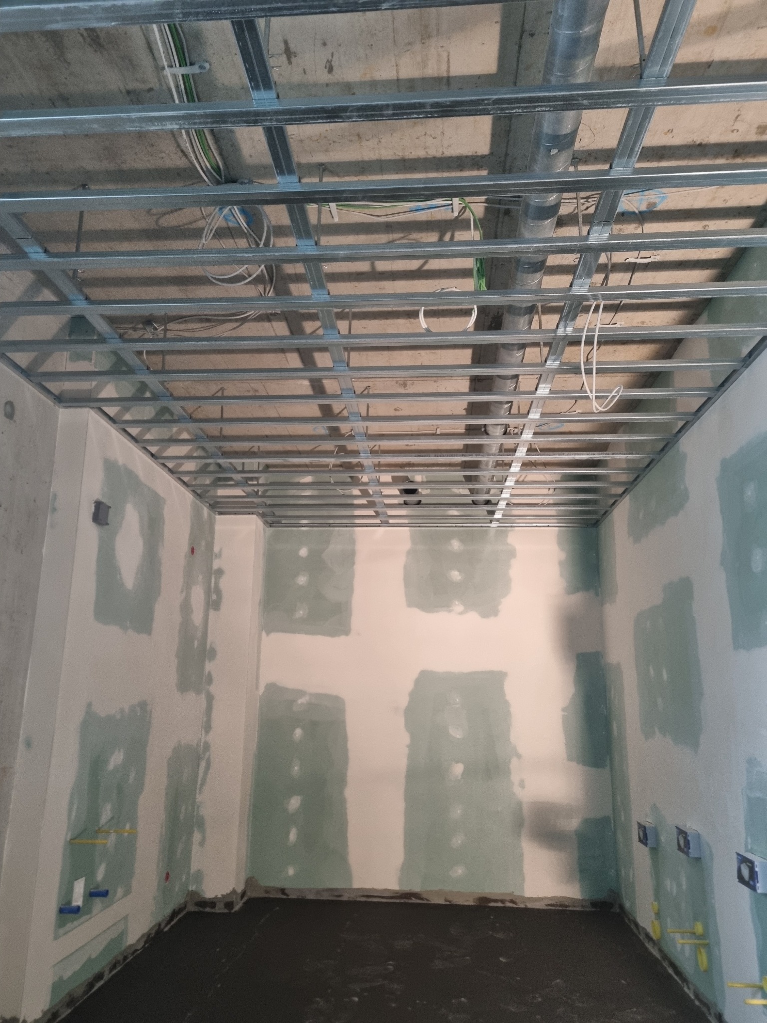 Montaż metalowej konstrukcji pod sufitem podwieszanym, widoczne przewody i instalacje, ściany w trakcie szpachlowania z widocznymi puszkami elektrycznymi.
