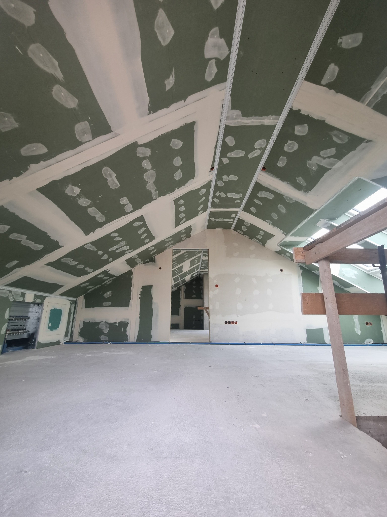 Poddasze w trakcie wykańczania płytami kartonowo-gipsowymi, widoczne szpachlowane łączenia i elementy konstrukcyjne, surowe ściany i podłoga, częściowo zamontowane instalacje.