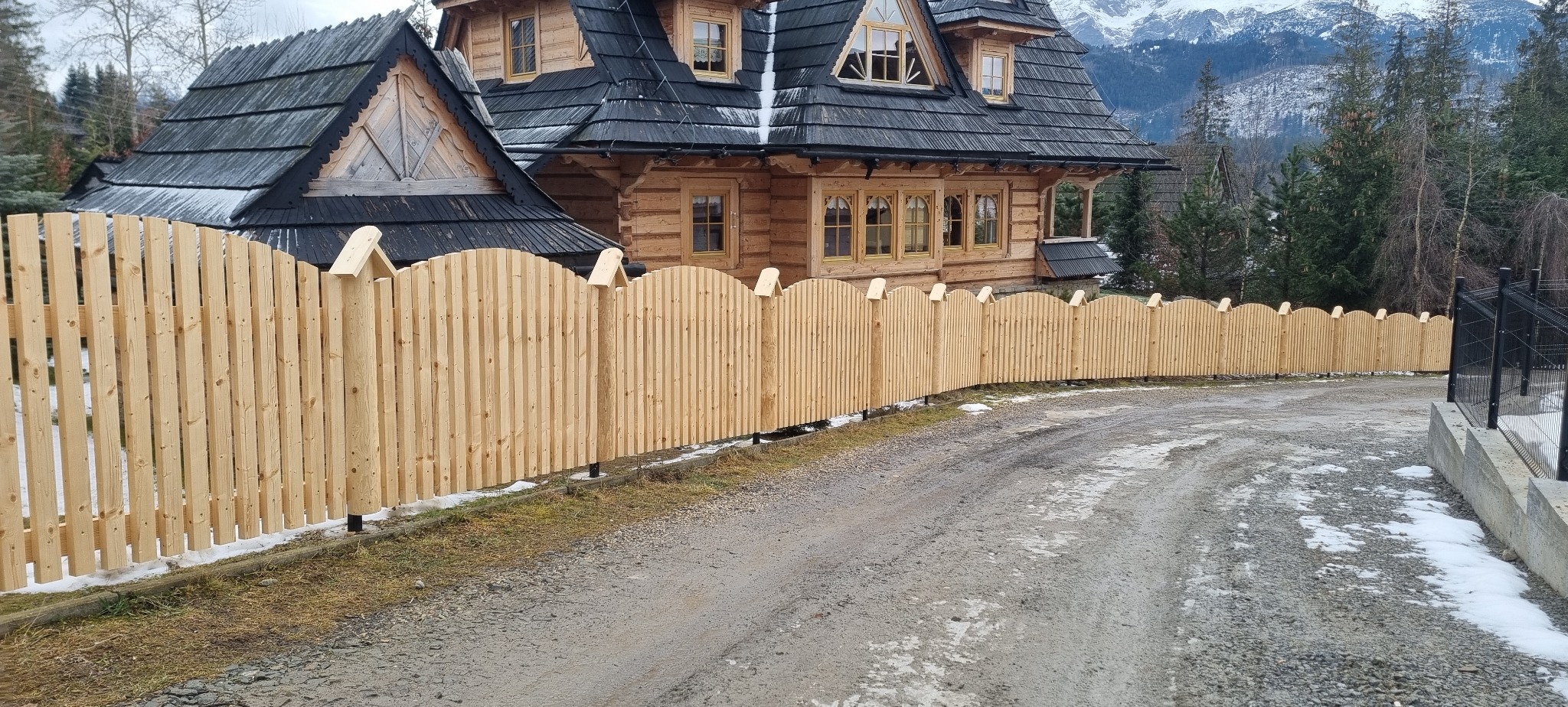 Nowe, jasne drewniane ogrodzenie sztachetowe z zaokrąglonymi szczytami, biegnące wzdłuż drogi gruntowej przed tradycyjnym domem góralskim z ciemnego drewna, w tle góry.