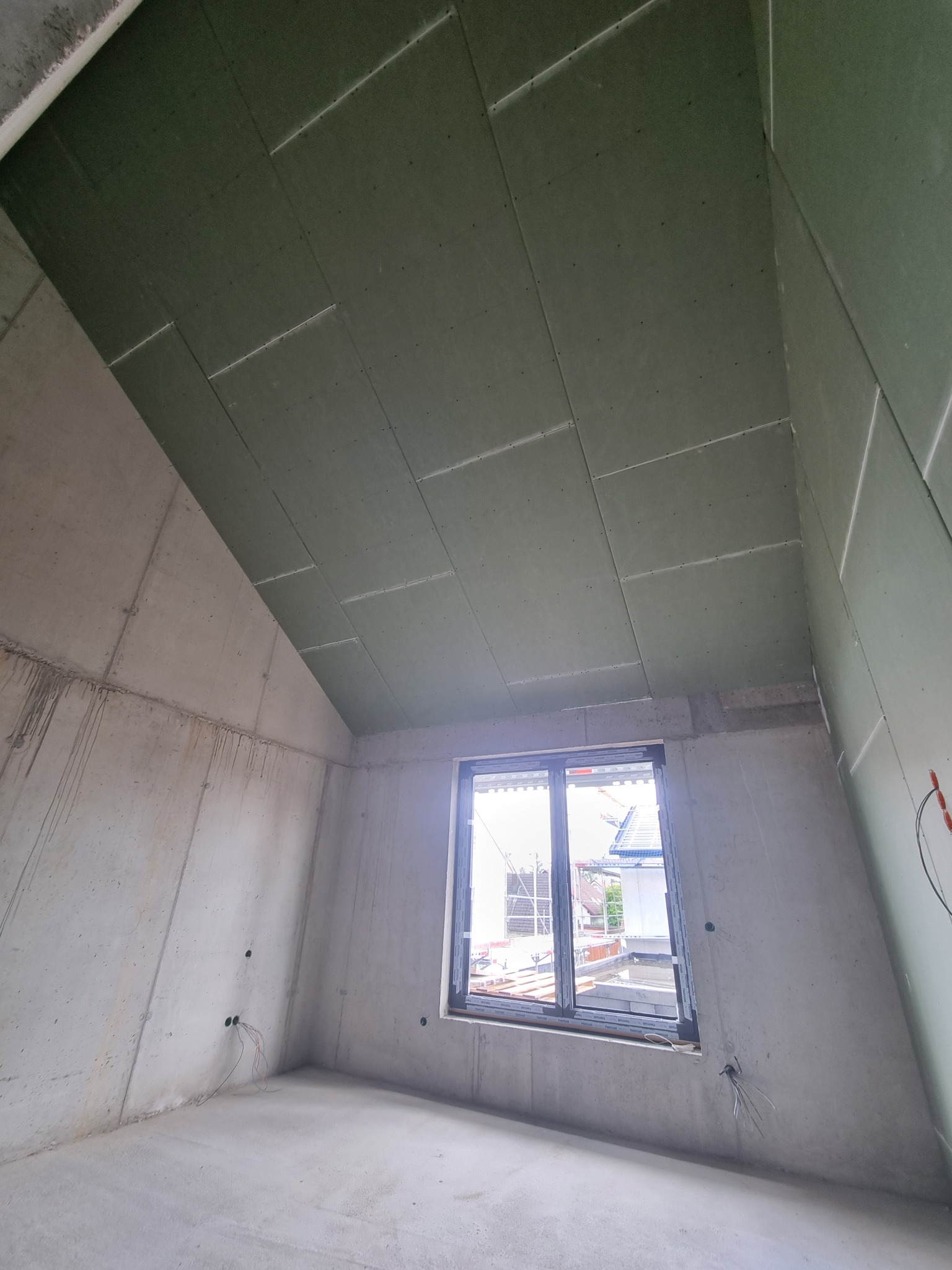 Wnętrze surowego pokoju na poddaszu z oknem, ściany z betonu, skośny sufit wykończony zielonymi płytami gipsowo-kartonowymi z widocznymi łączeniami.